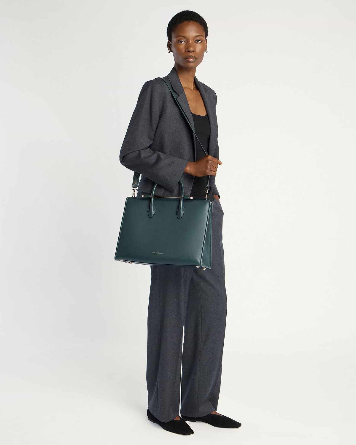 Woman in grey suit standing, holding Strathberry bottle green tote bag with strap.