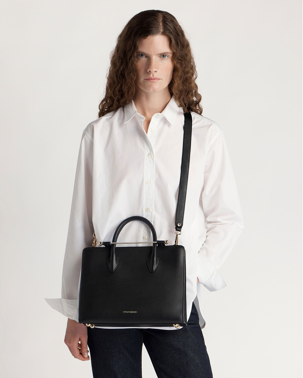 Woman wearing white shirt carrying black Strathberry midi tote with top handles and shoulder strap.