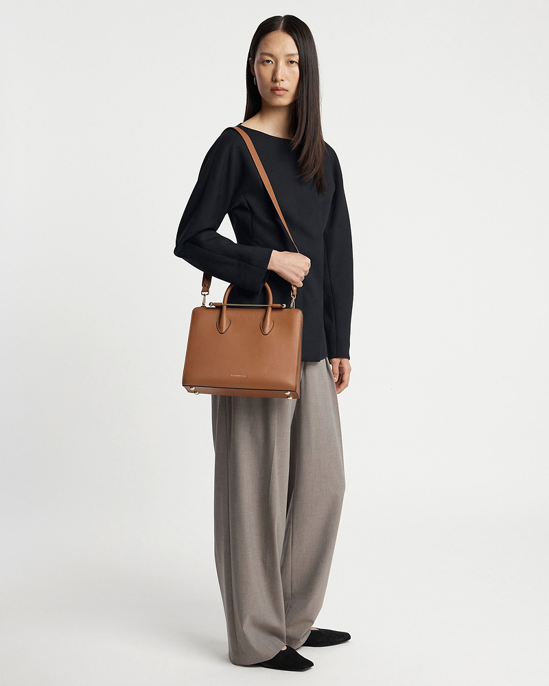 Model wearing black top and grey wide trousers holding chestnut midi tote bag.