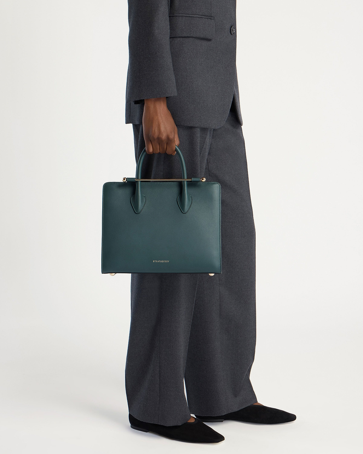 Model wearing grey suit holding bottle green Strathberry midi tote at side