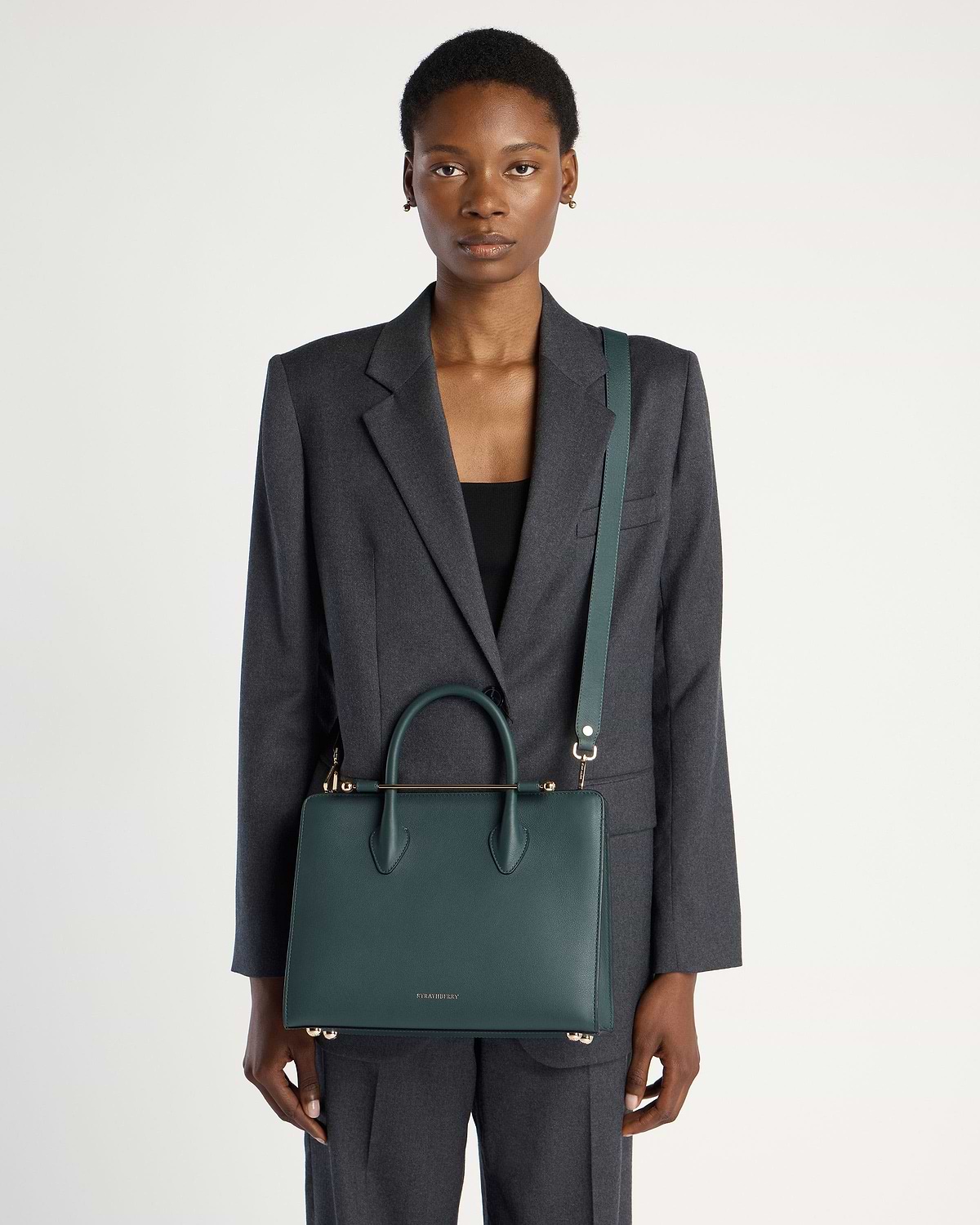 Model wearing grey suit carries bottle green Strathberry midi tote with shoulder strap