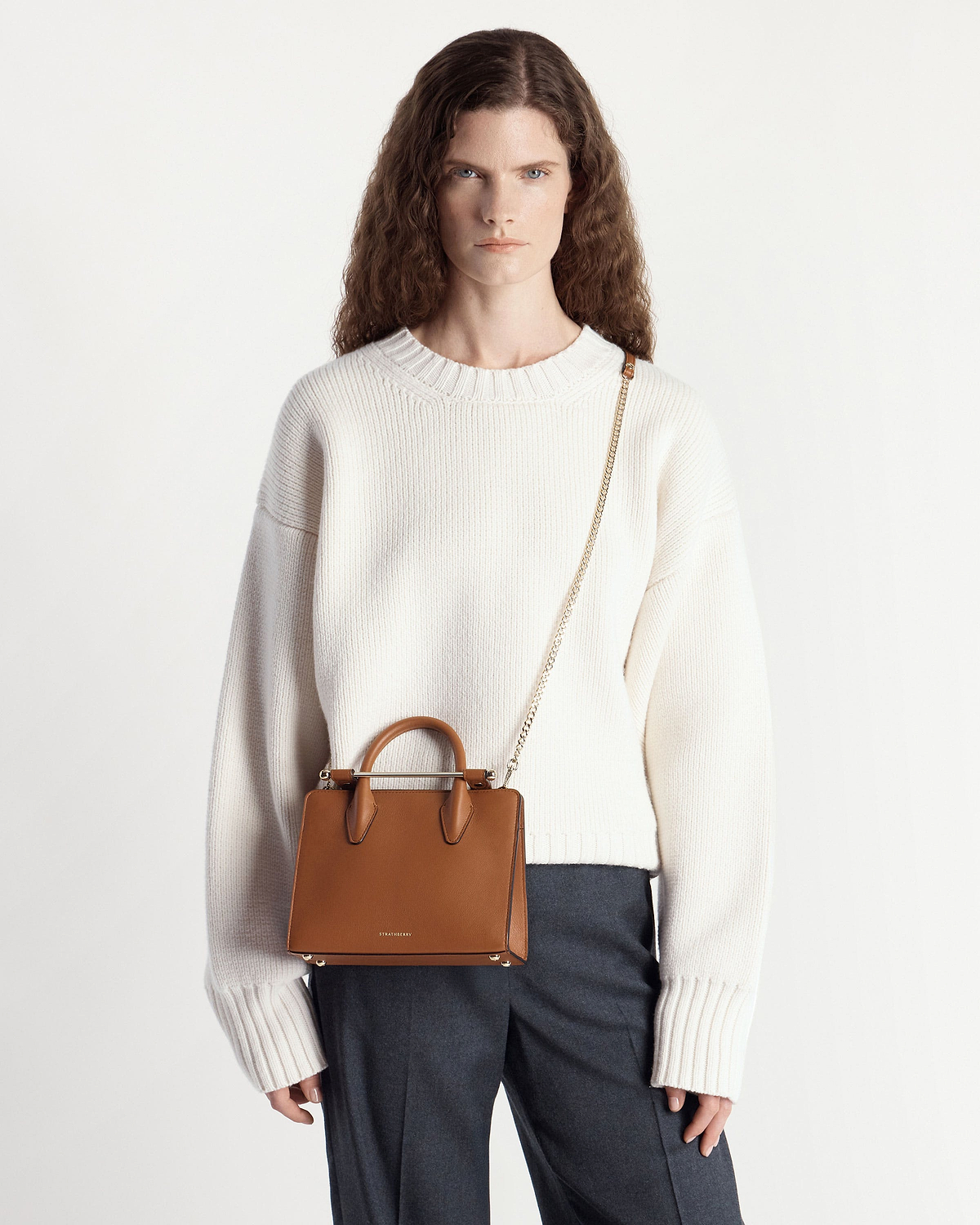 Model wearing chestnut Strathberry mini tote with chain strap over cream jumper.