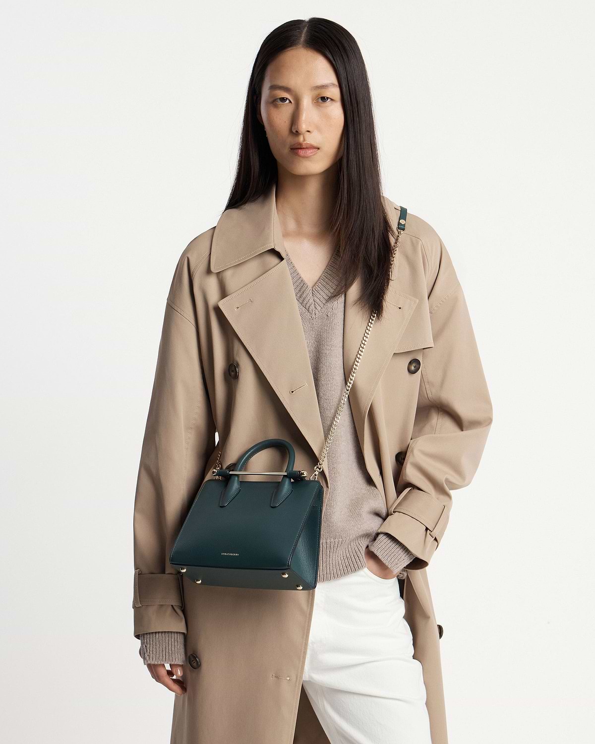 Model wearing beige trench carrying bottle green Strathberry mini tote with chain strap
