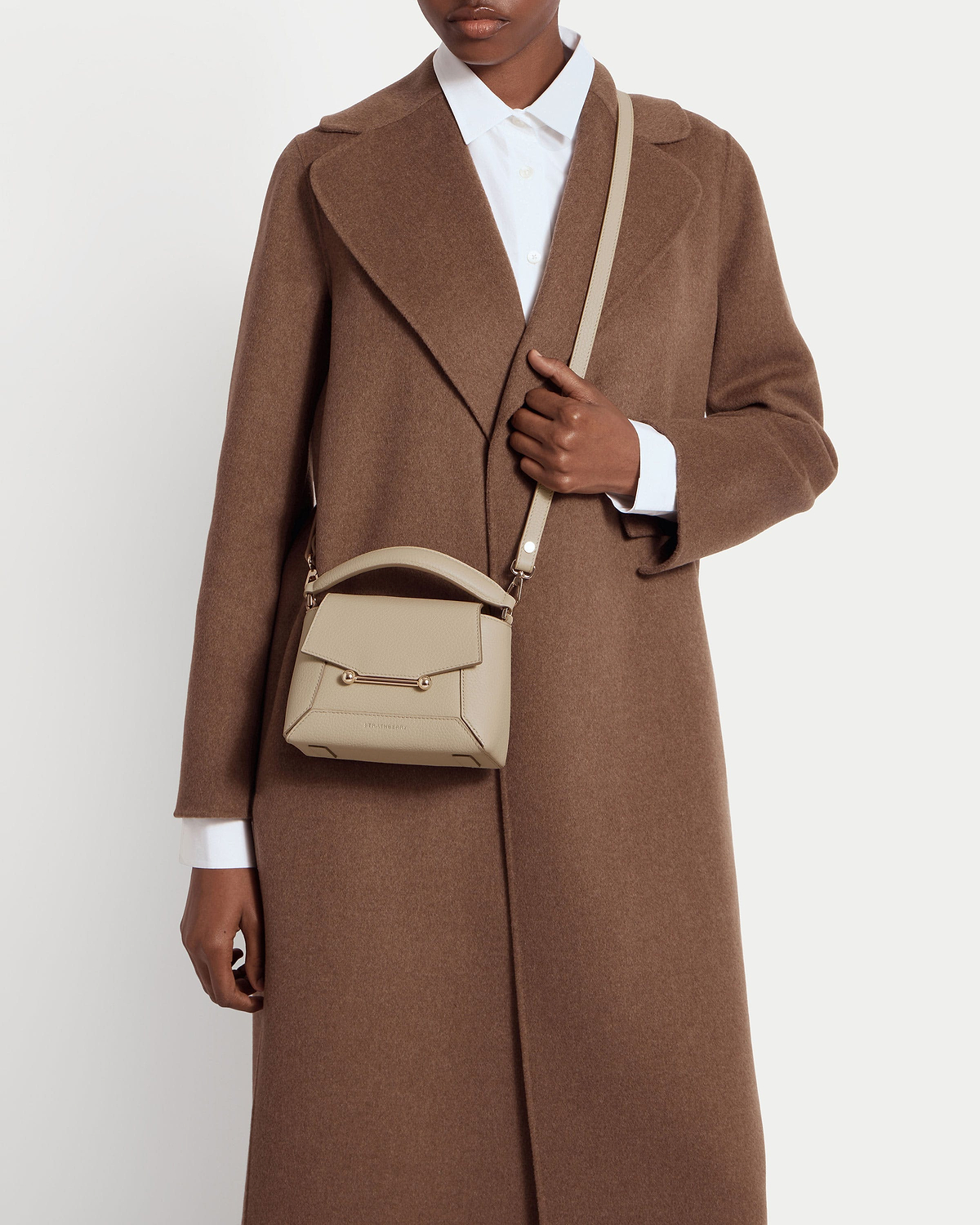Model in brown coat holds an oat-coloured small leather top-handle crossbody bag.
