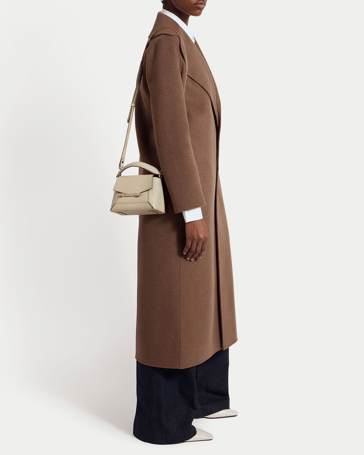 Model wearing a long brown coat with an oat Mosaic Nano shoulder bag.