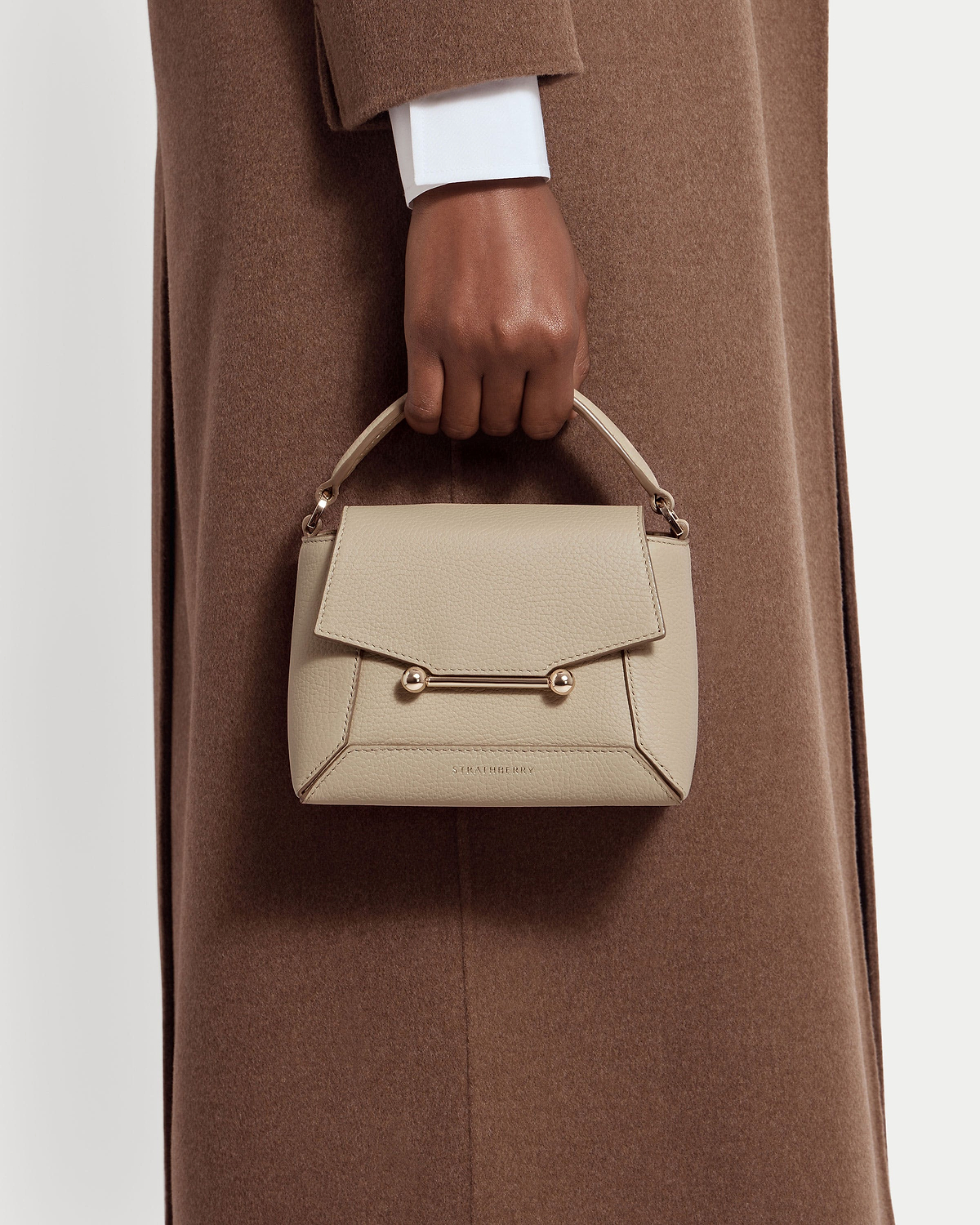 Hand holding Strathberry Mosaic Nano top-handle bag in oat leather against brown coat.