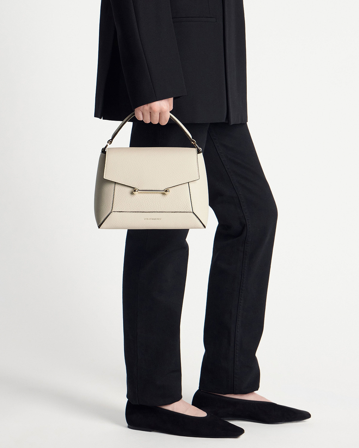 Model holding a vanilla Strathberry Mosaic bag by its handle, wearing black trousers and flats.