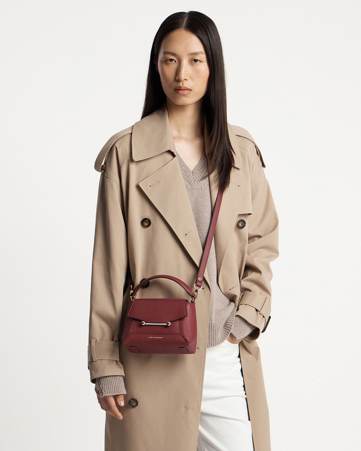 Model wearing beige trench coat carrying Strathberry Mosaic Nano bag in oxblood crossbody.