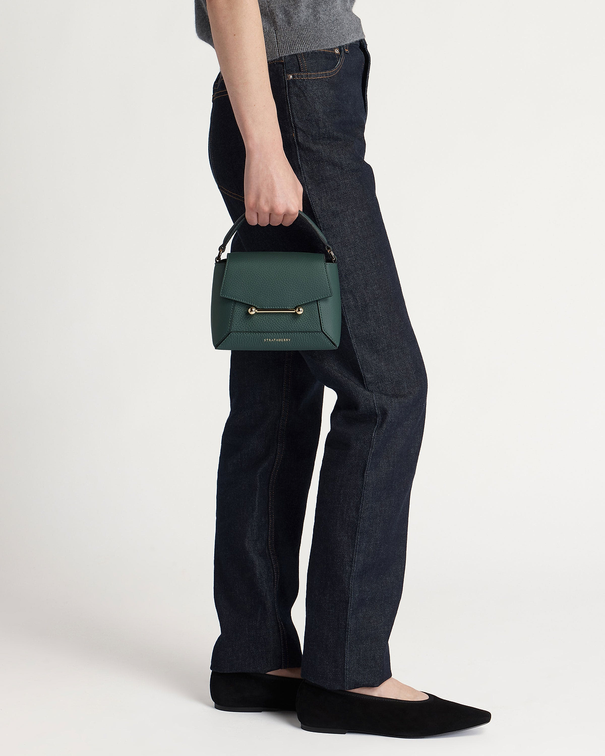Model holding bottle green Strathberry leather top-handle handbag by her side.