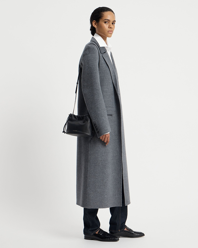 Model in grey coat carrying a black drawstring shoulder bag at her side.