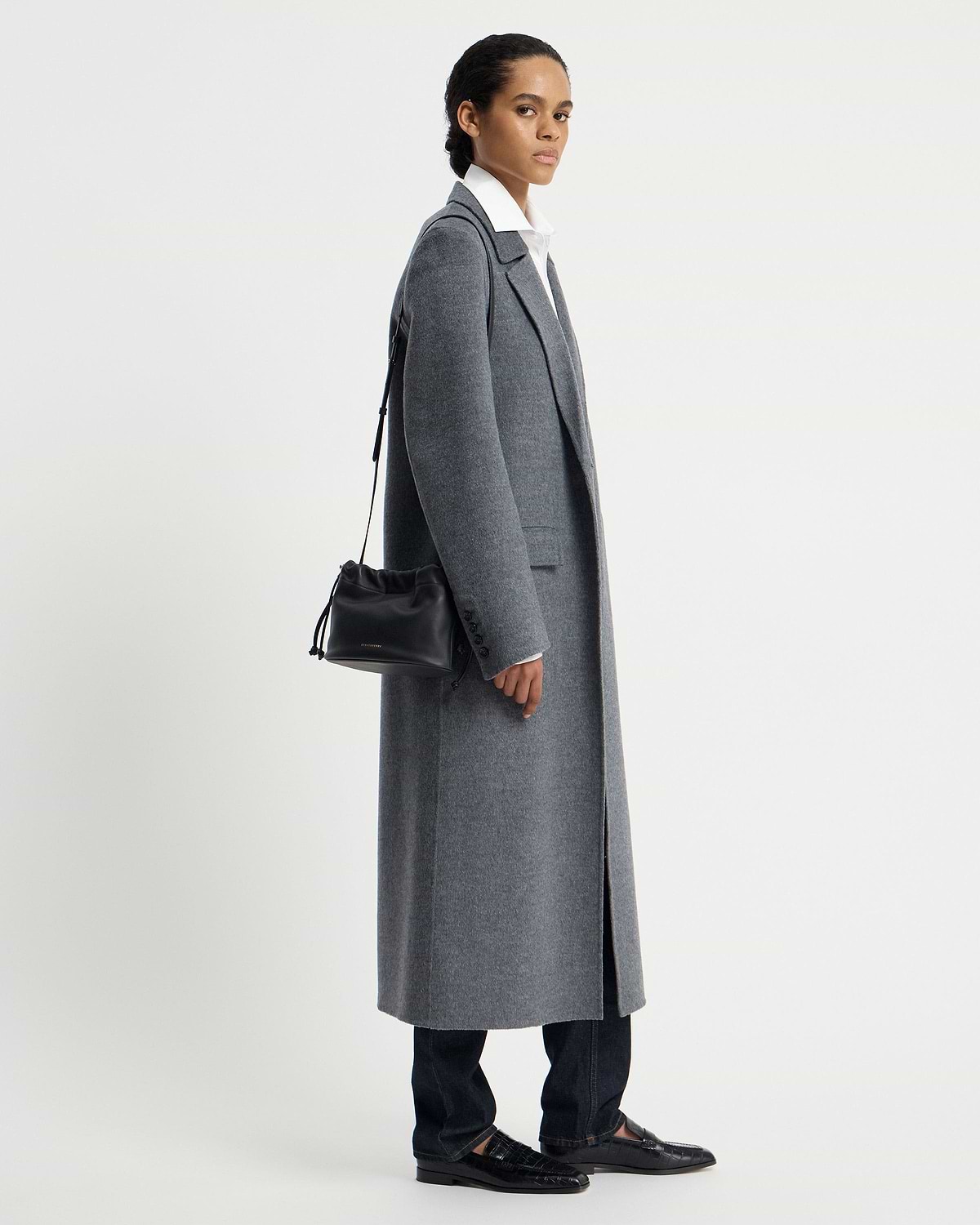 Model in grey coat carrying a black drawstring shoulder bag at her side.