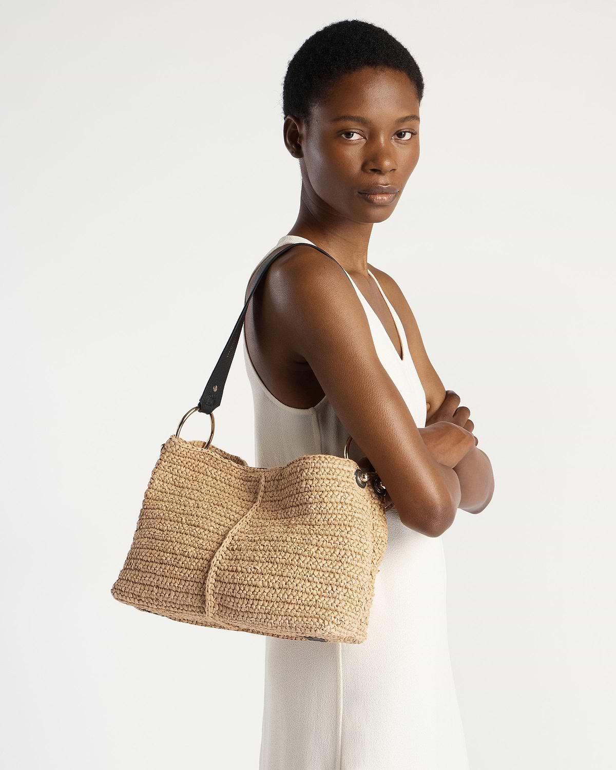 Model wearing white sleeveless dress carrying raffia hobo bag with black strap.