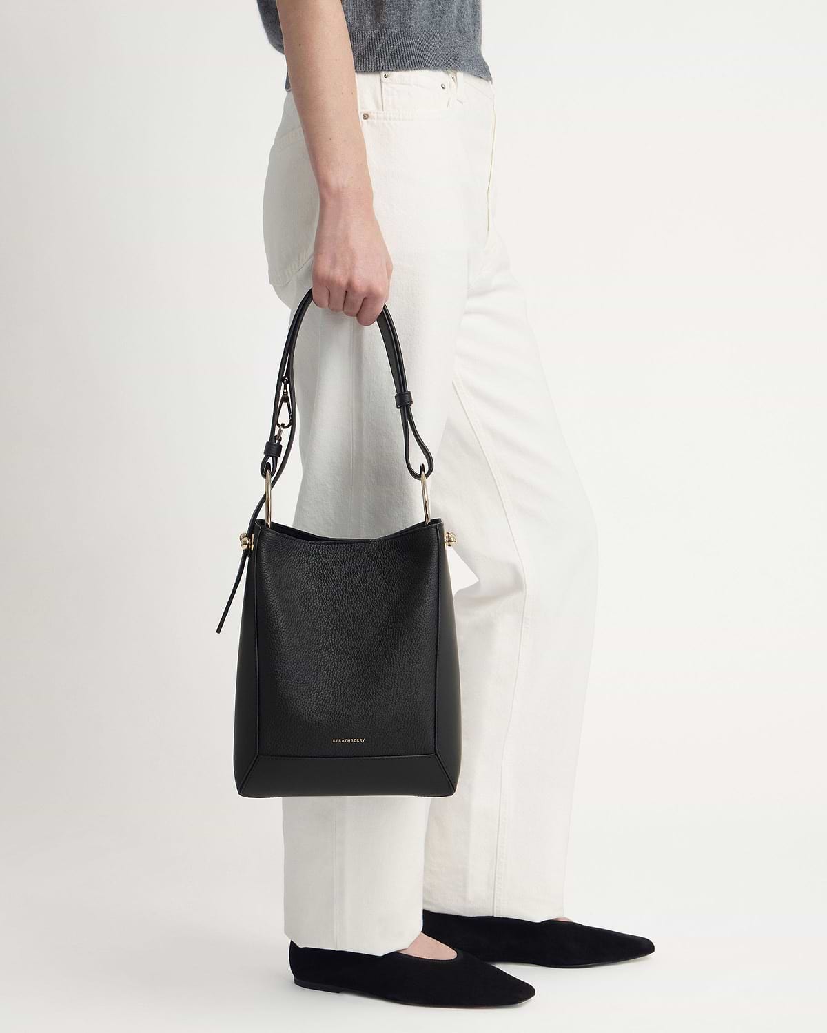 Model holding black Strathberry bucket bag with gold hardware and white trousers.