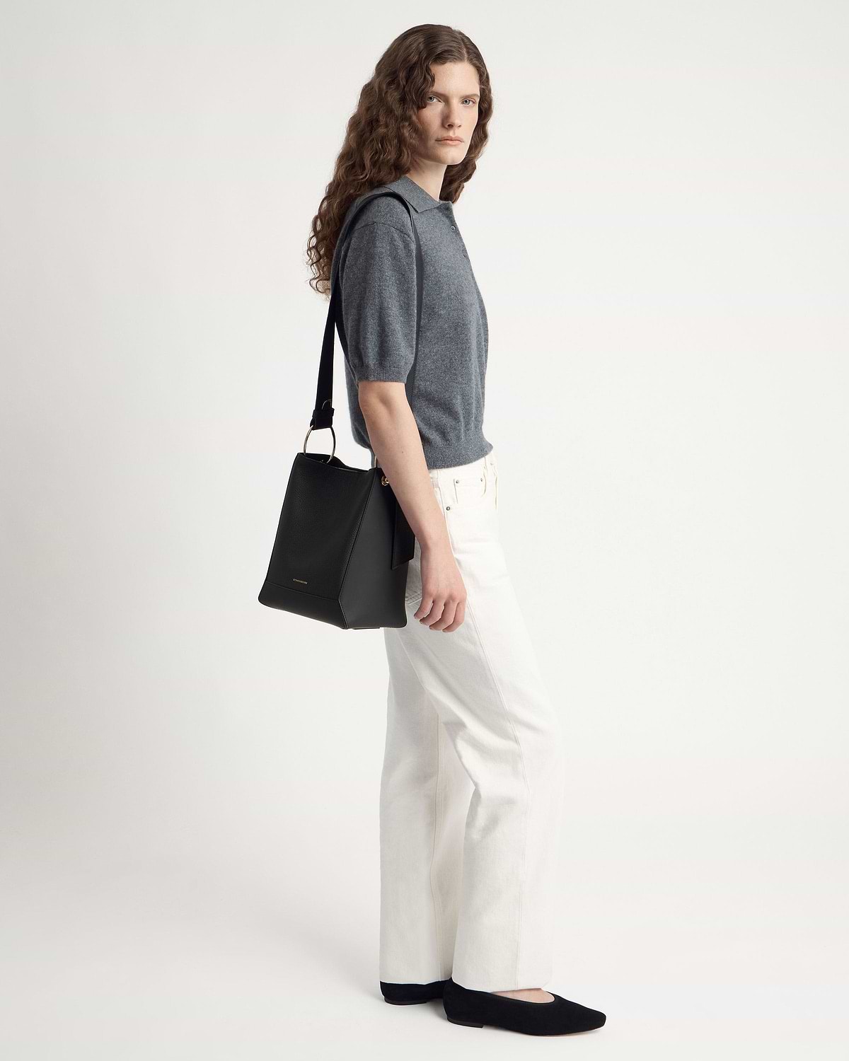Model wearing grey short-sleeve polo and white jeans carrying black bucket bag over shoulder