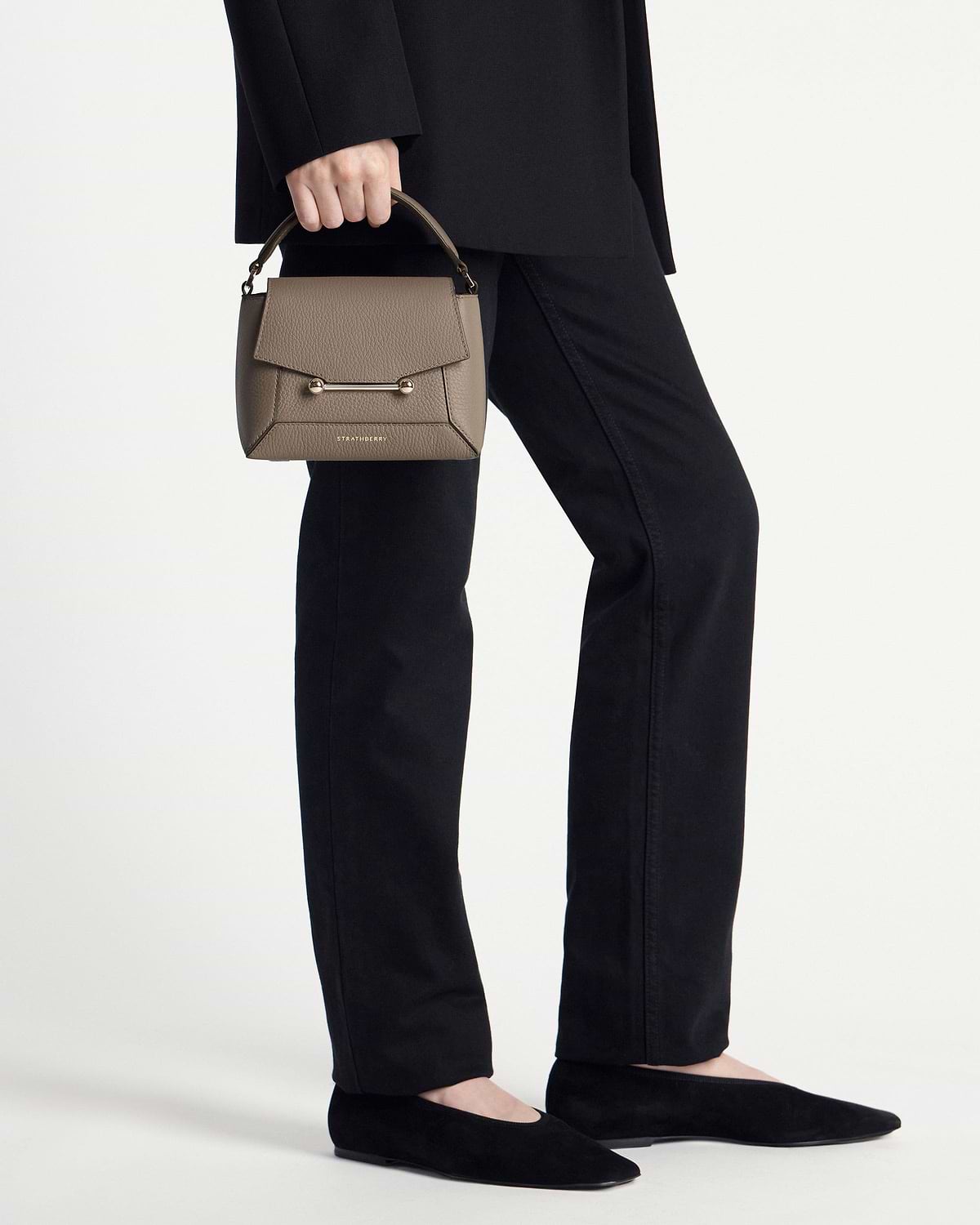 Model waist-down holding taupe Strathberry Mosaic Nano top-handle bag with gold bar closure.