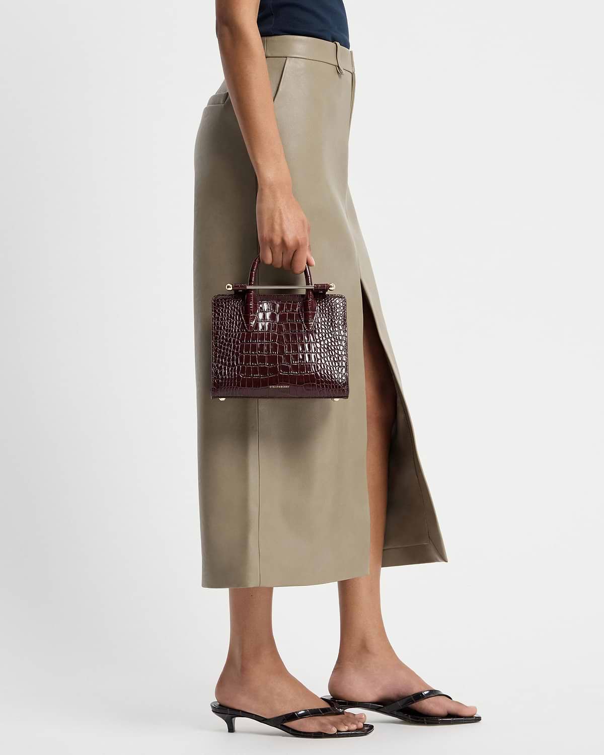 Model's hand holding burgundy croc mini tote by Strathberry beside skirt.