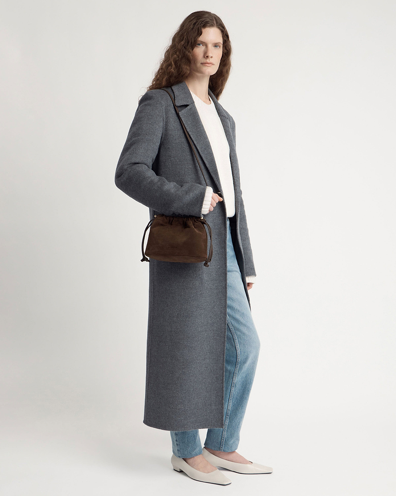 Model wearing long grey coat and jeans carrying chocolate suede drawstring bag.