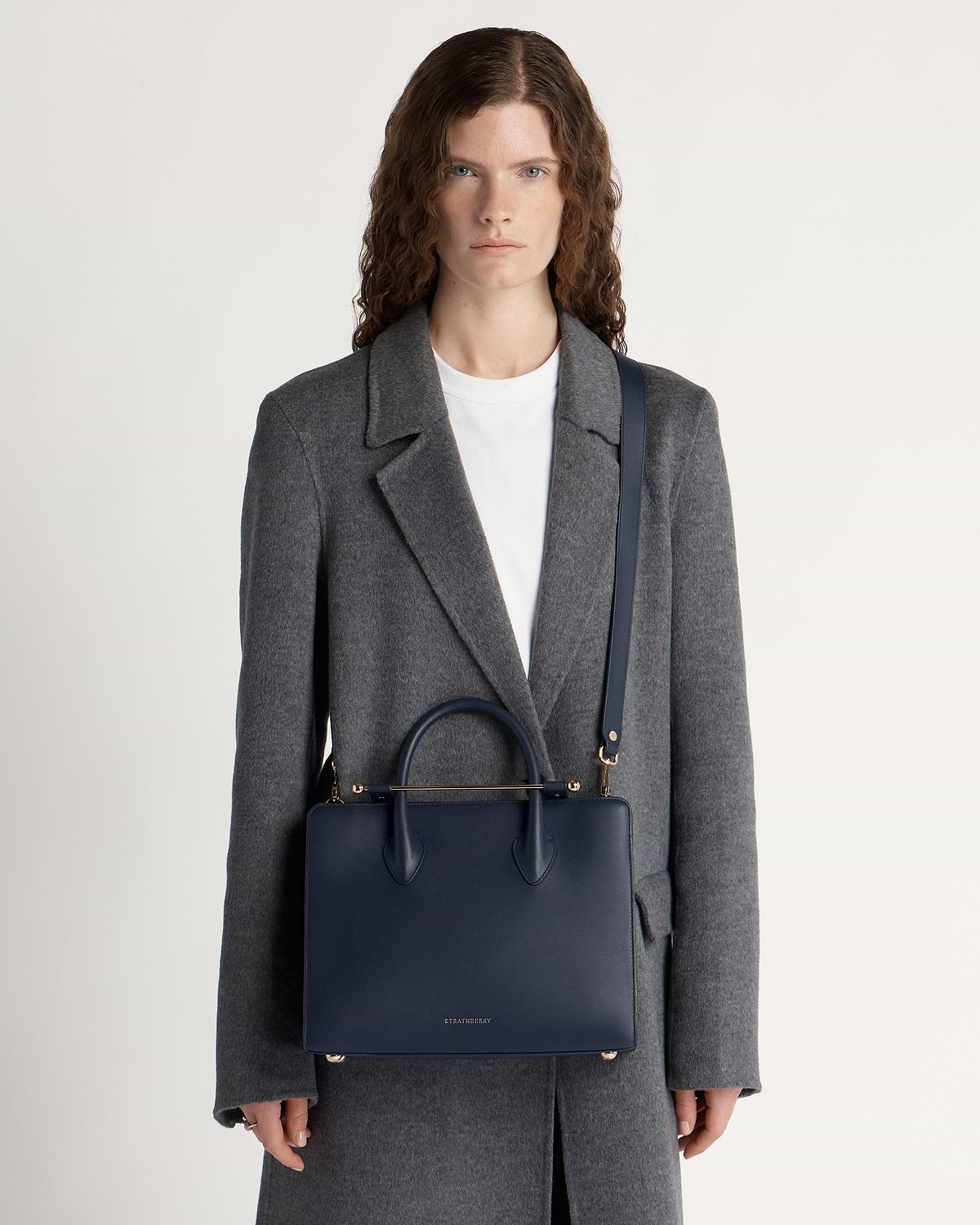 Model in grey coat carrying marine blue Strathberry midi tote with shoulder strap.