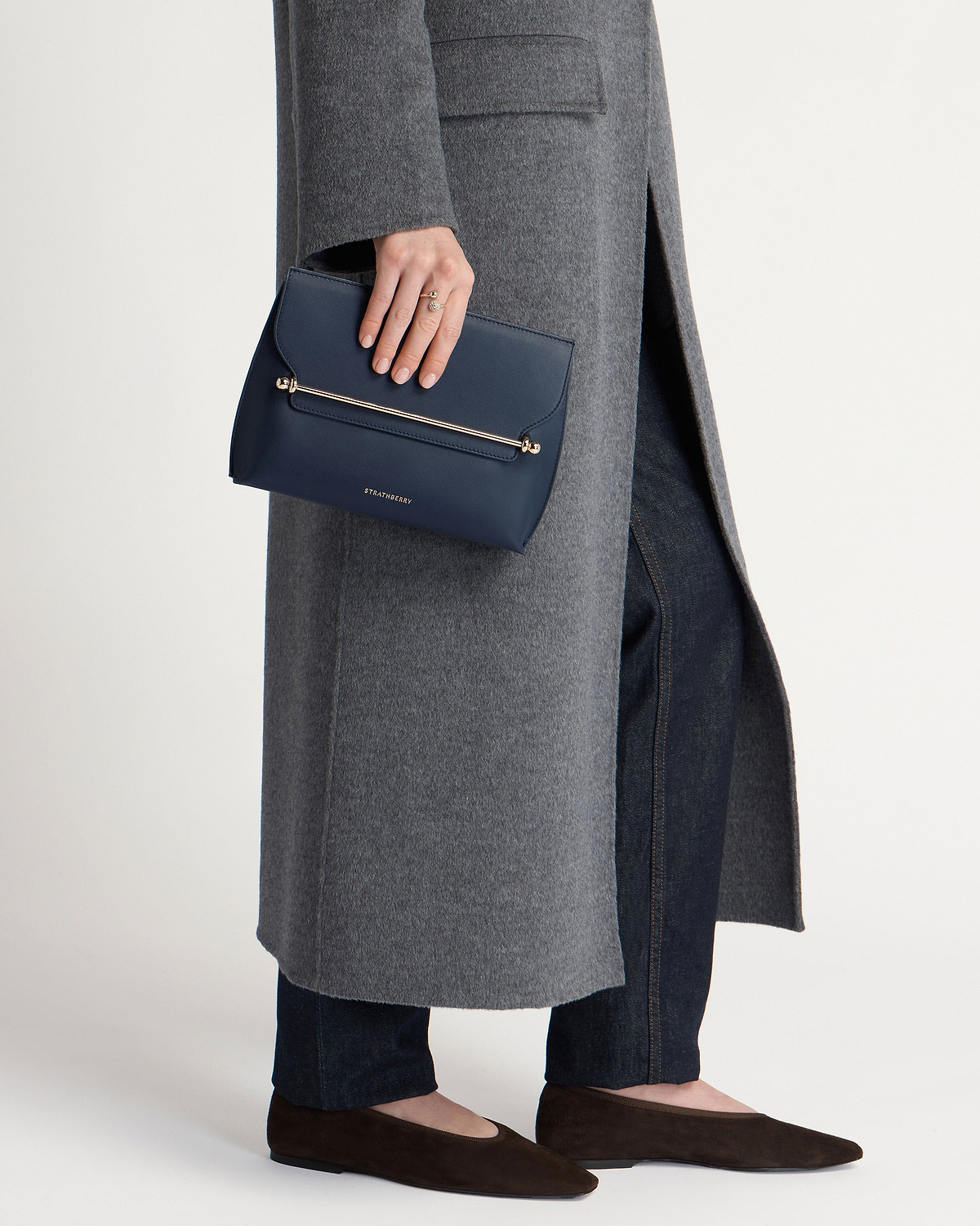 Hand holding marine blue Strathberry Stylist clutch with gold bar, grey coat.