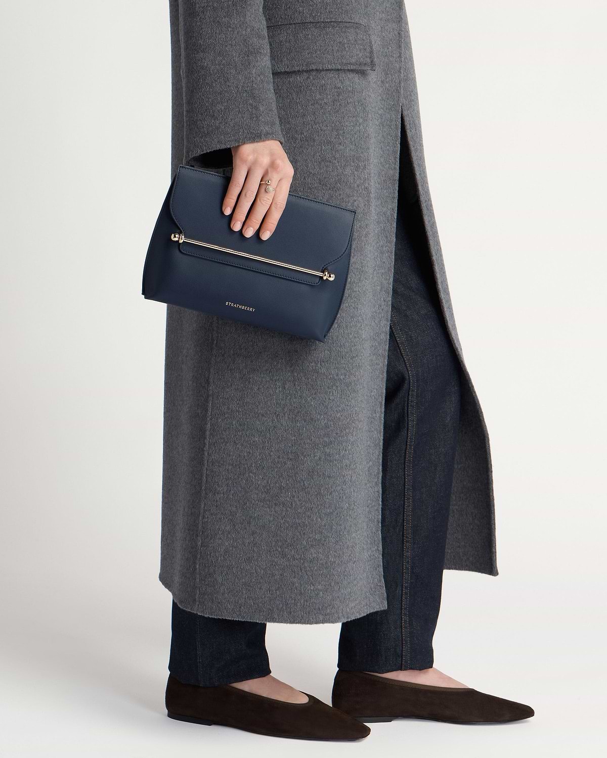 Hand holding marine blue Strathberry Stylist clutch with gold bar, grey coat.