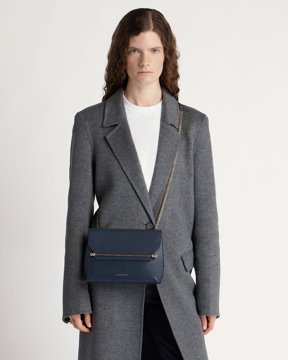 Model wearing marine blue Strathberry Stylist bag with gold bar and chain strap.