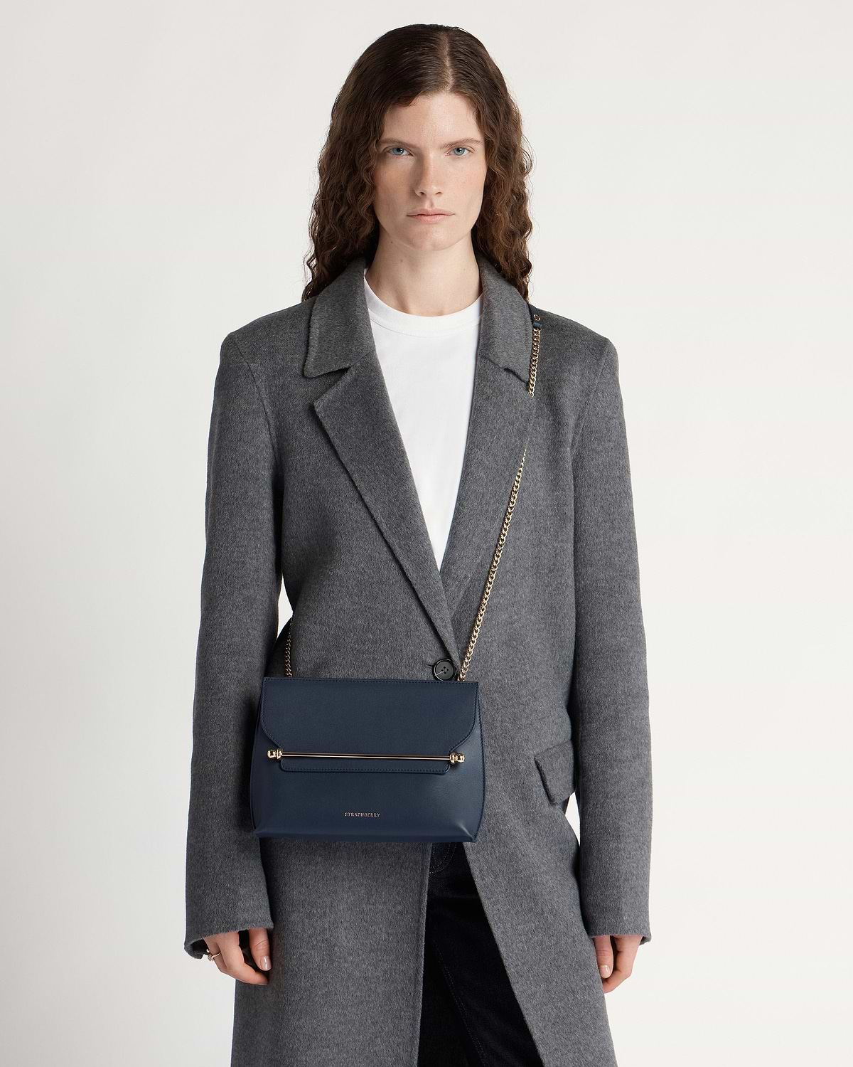 Model wearing marine blue Strathberry Stylist bag with gold bar and chain strap.