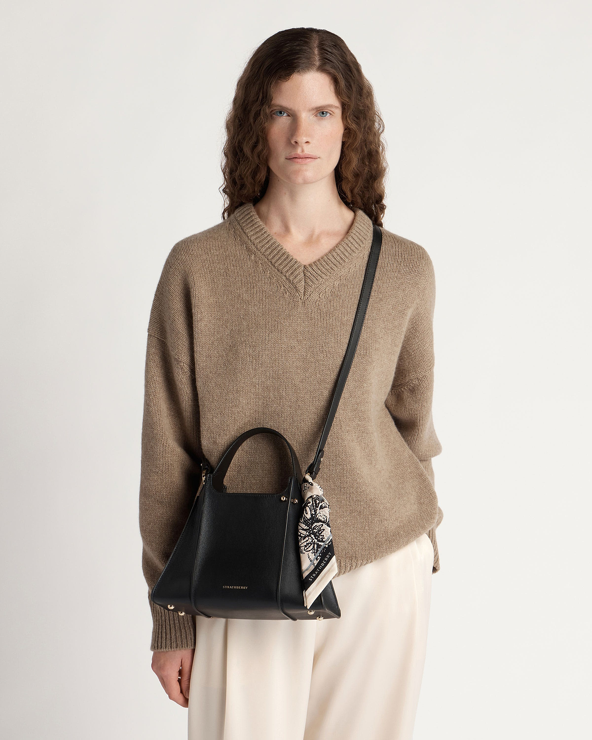 Model wearing beige jumper carrying black Strathberry Arkshell bag with shoulder strap and scarf.