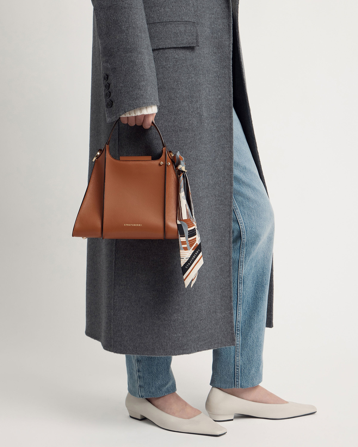 Person holding tan Strathberry Arkshell handbag with patterned scarf, grey coat and jeans.