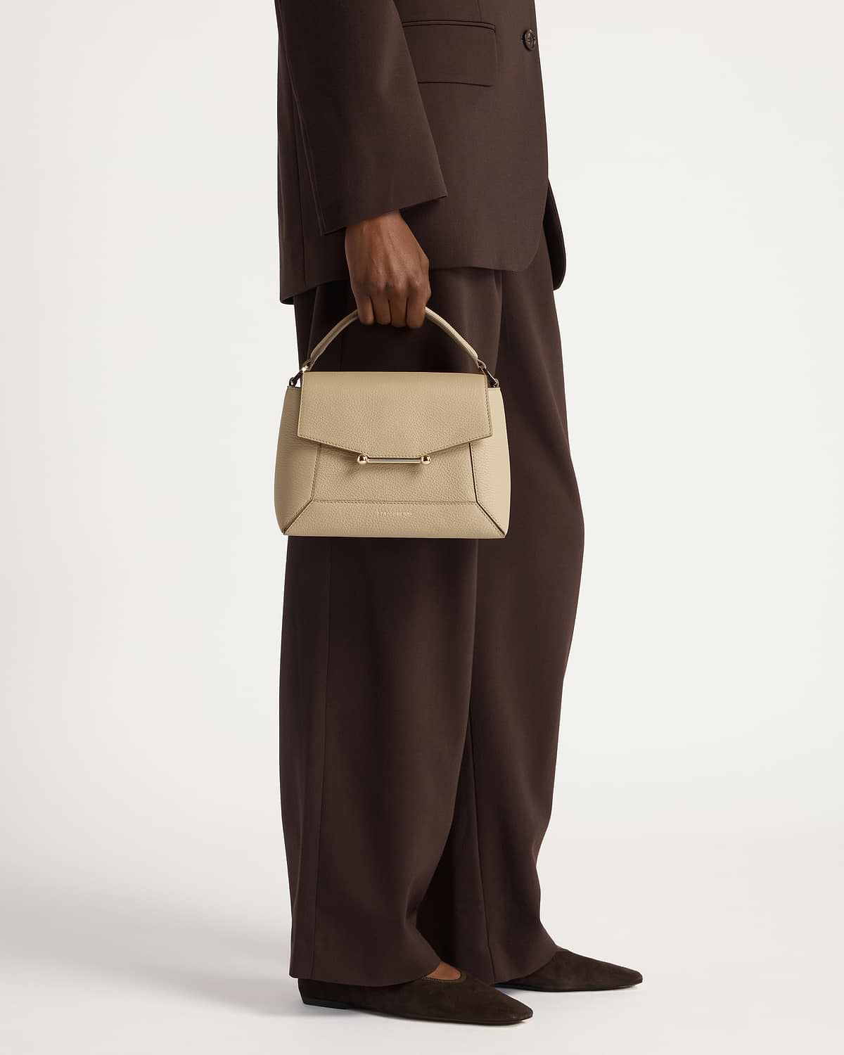 Model holding a sand mosaic top-handle bag against a brown tailored suit.