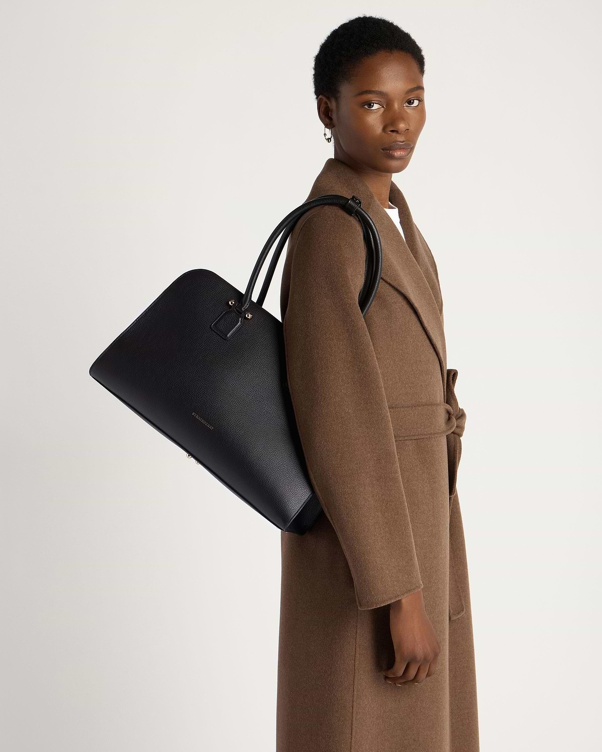 Model in brown coat carrying black Strathberry Georgia Maxi leather tote on shoulder