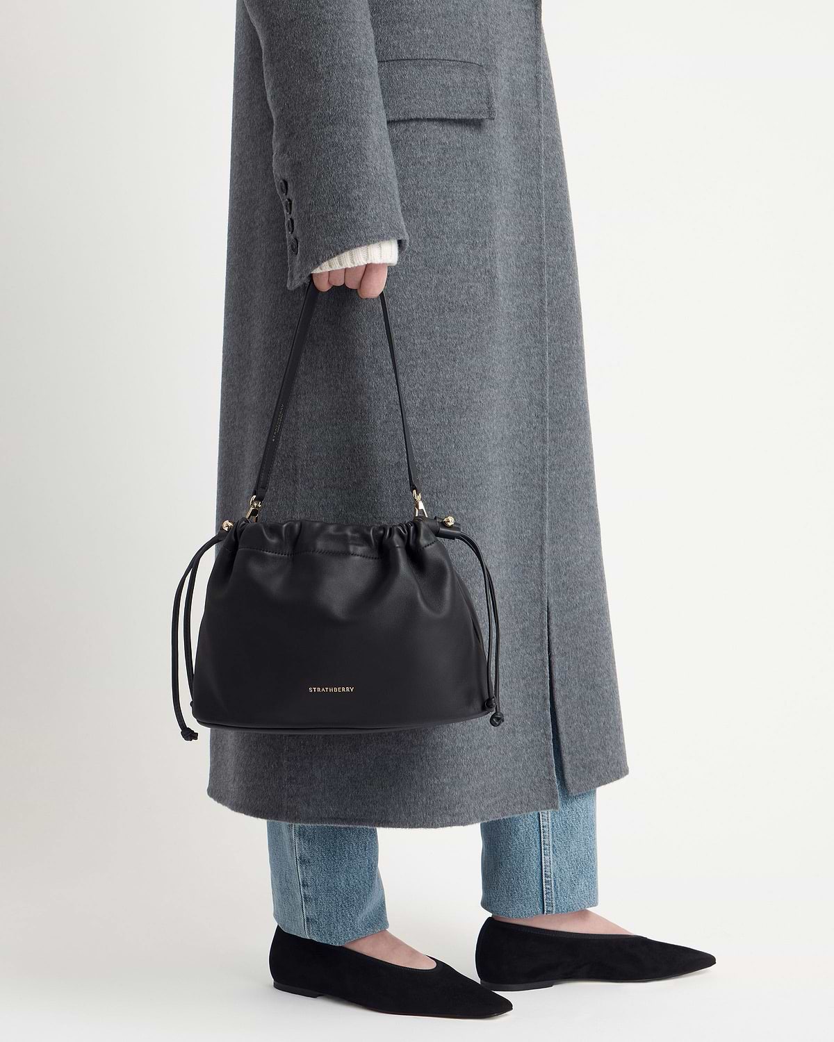 Black Strathberry Charlotte Midi drawstring bag held by model wearing grey coat.