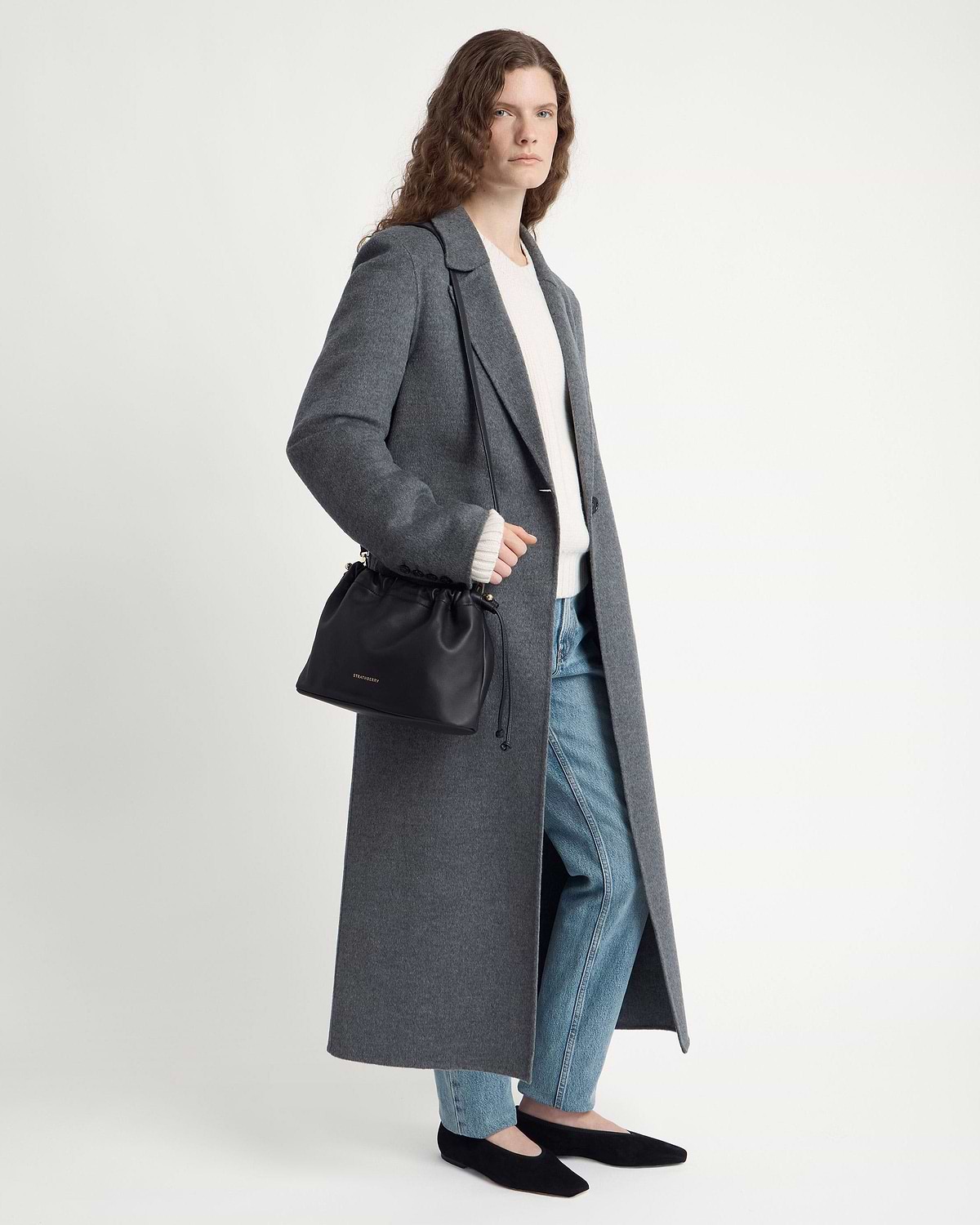Model wearing a grey long coat and jeans carries a Strathberry black drawstring bag.