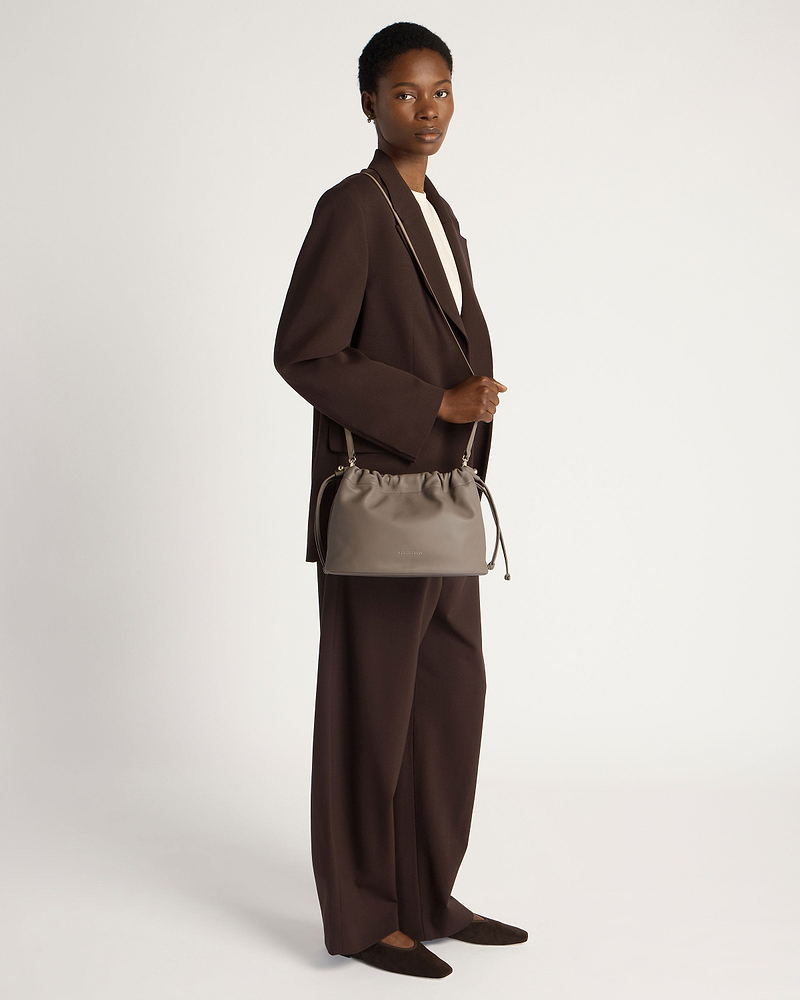 Model in brown suit holding a taupe drawstring shoulder bag at her side.