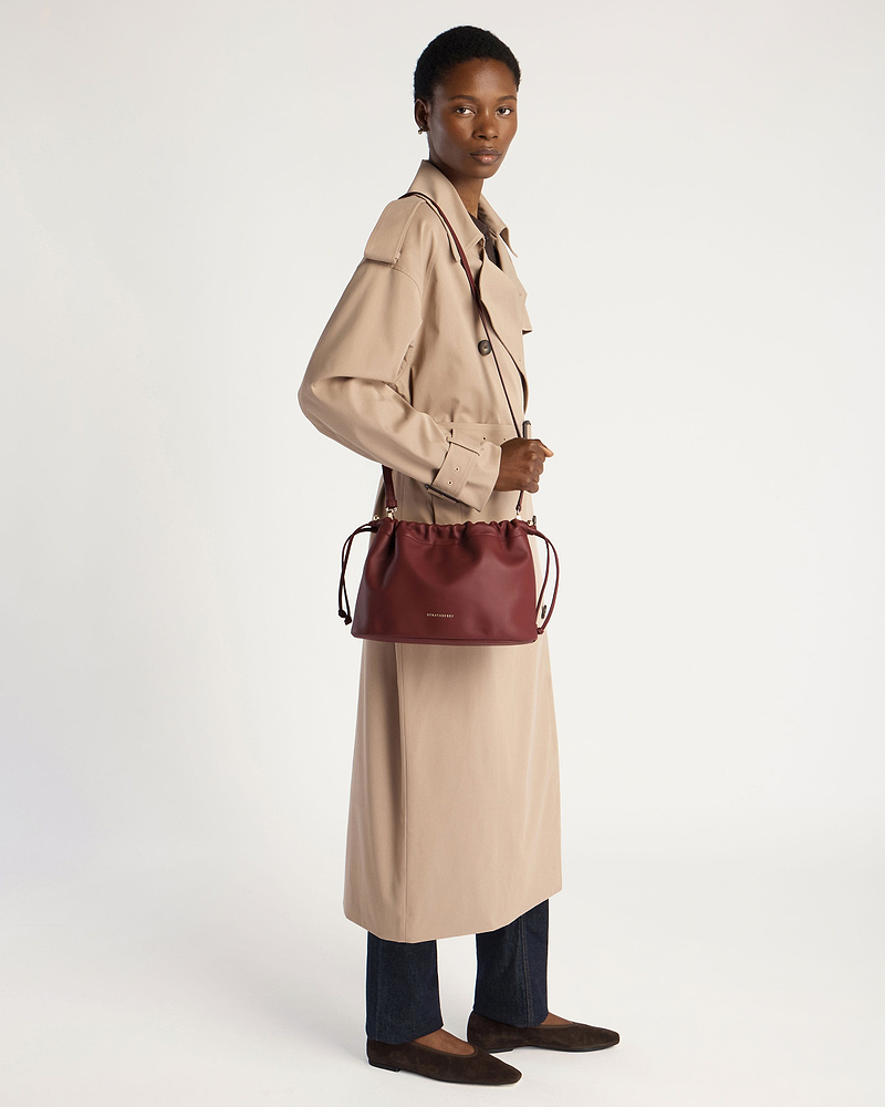 Model in beige trench coat holding oxblood Strathberry drawstring bag over shoulder.