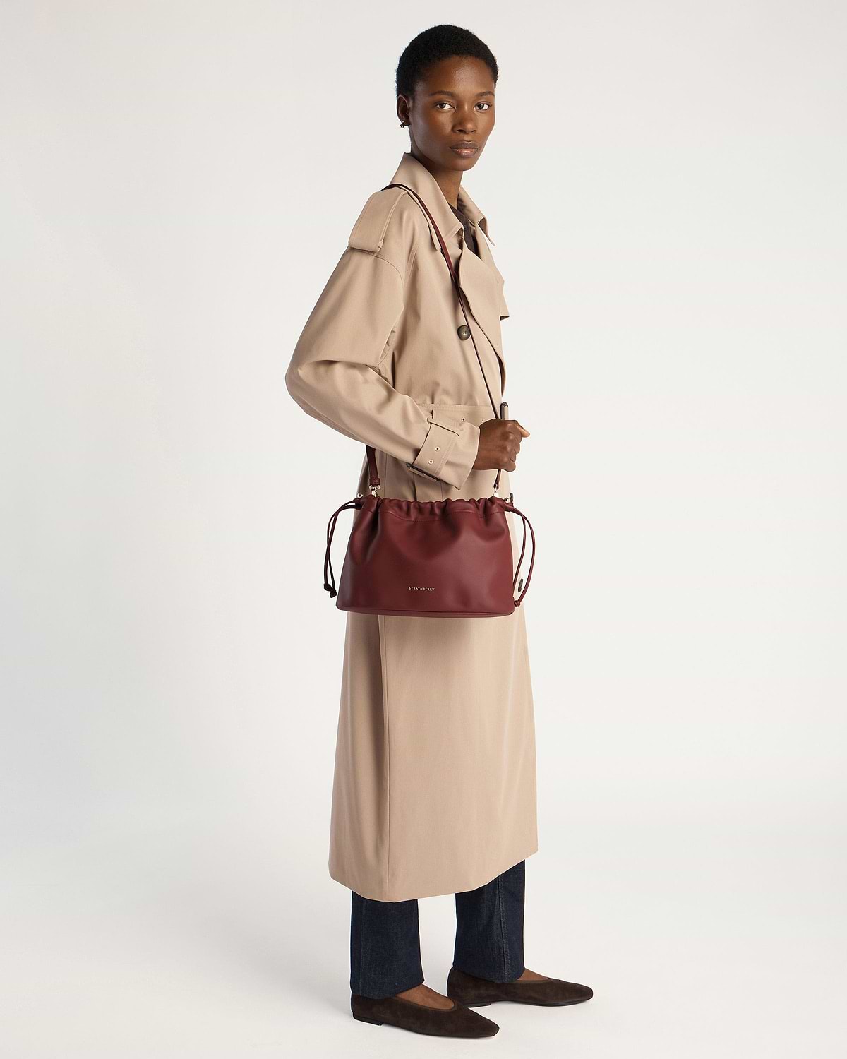 Model in beige trench coat holding oxblood Strathberry drawstring bag over shoulder.