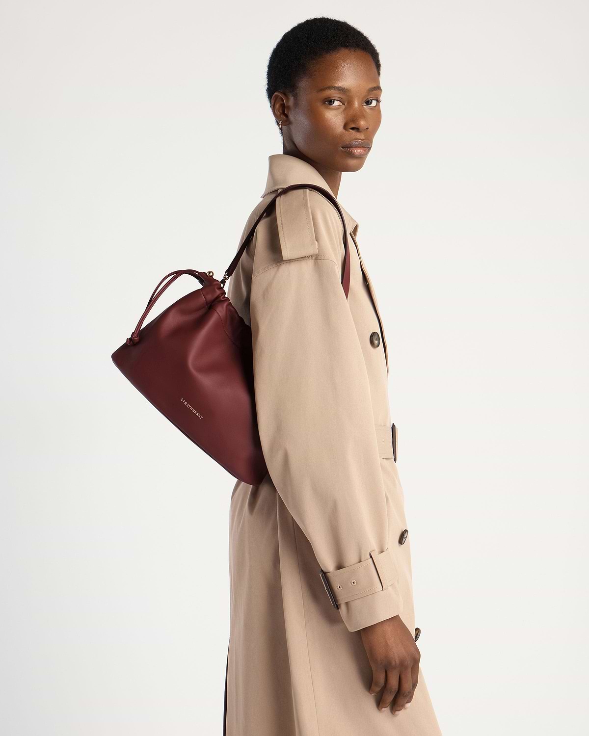 Model wearing a beige trench coat carrying oxblood Strathberry drawstring bag.