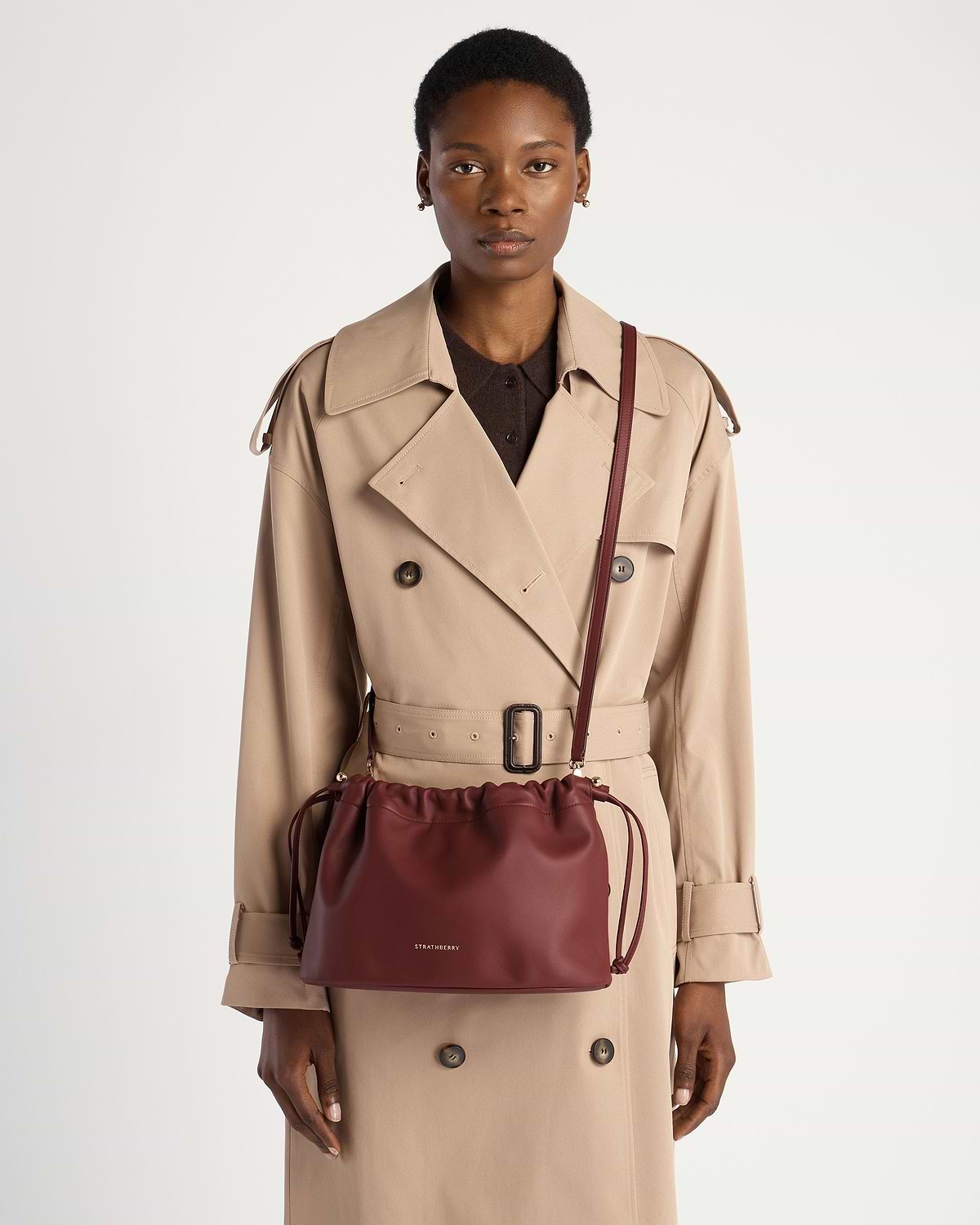 Model wearing beige trench coat carrying oxblood drawstring crossbody bag with Strathberry logo.