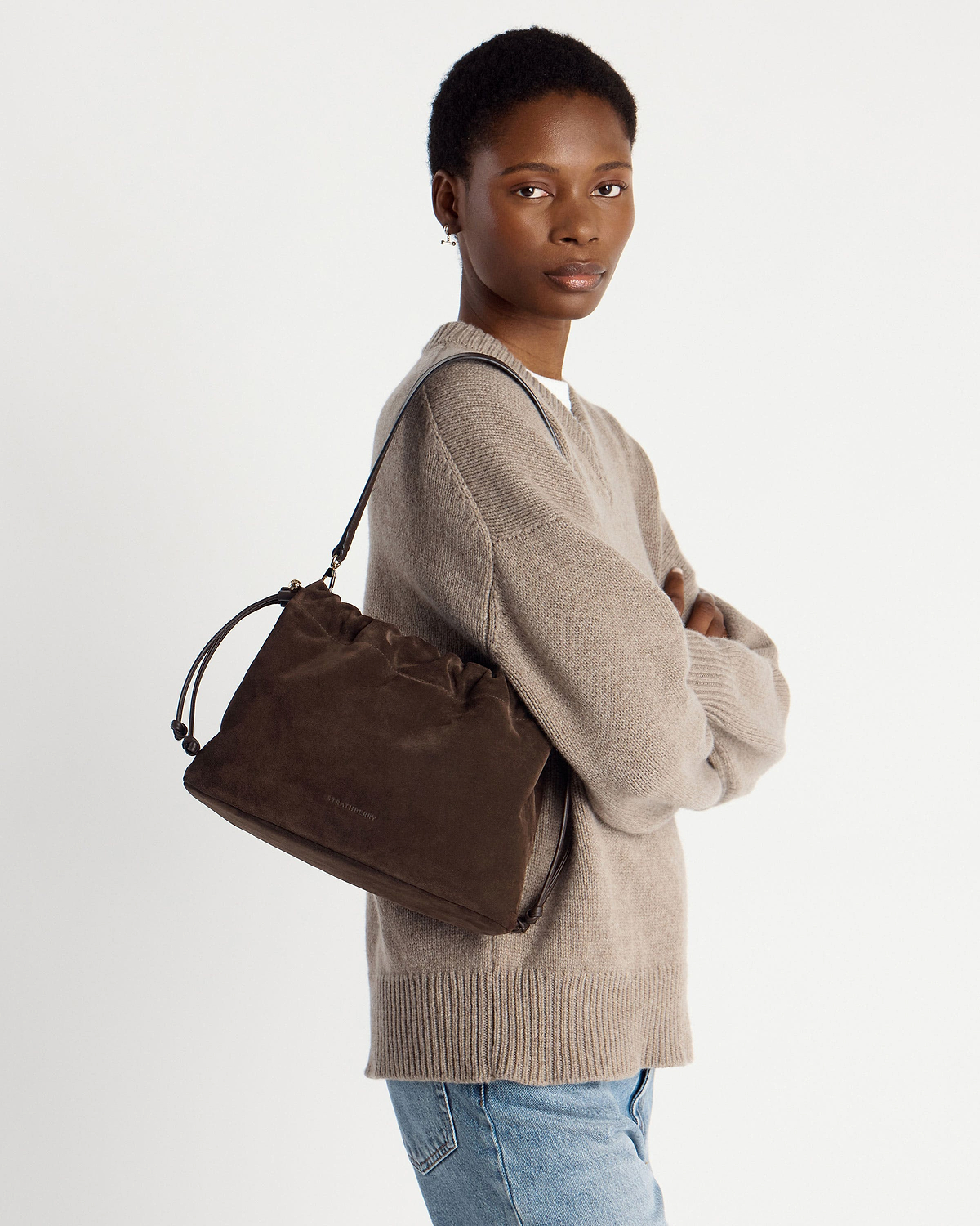 Model wearing a chocolate suede drawstring bag over shoulder, arms crossed.