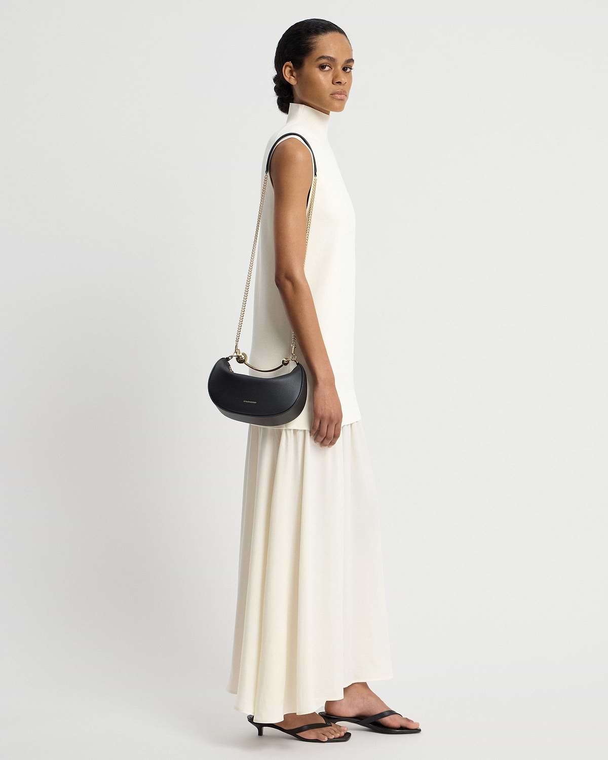 Model in cream dress carrying black crescent-shaped mini shoulder bag with gold chain.