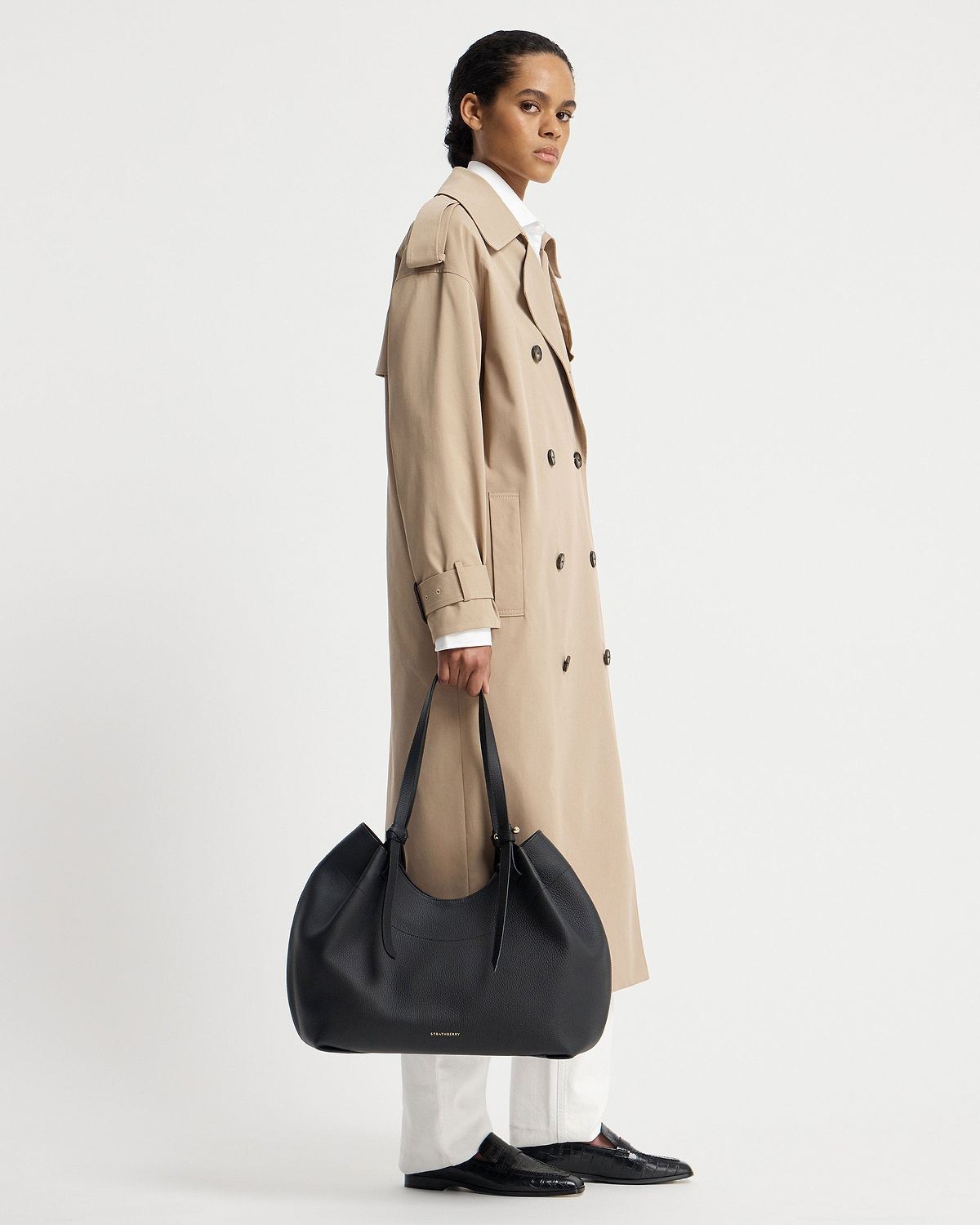 Model in a beige trench coat holding a black Strathberry Barra Tote at her side.