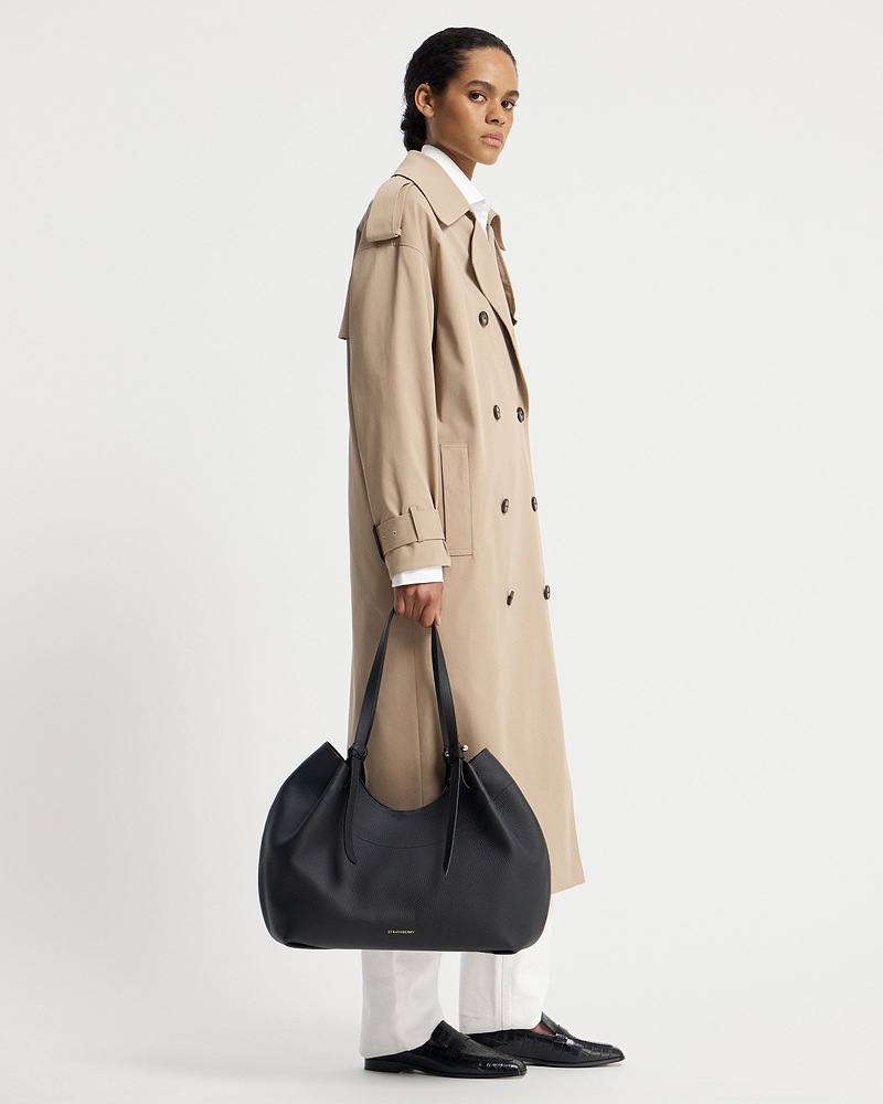 Model in a beige trench coat holding a black Strathberry Barra Tote at her side.