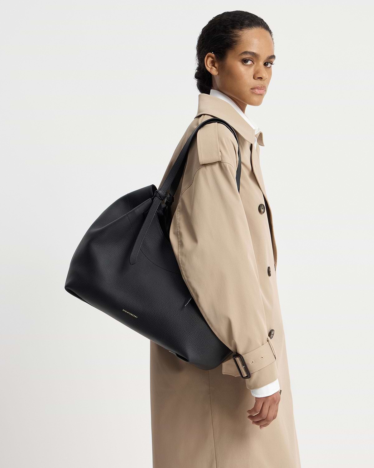Model in a beige trench coat carrying a black Strathberry Barra tote.