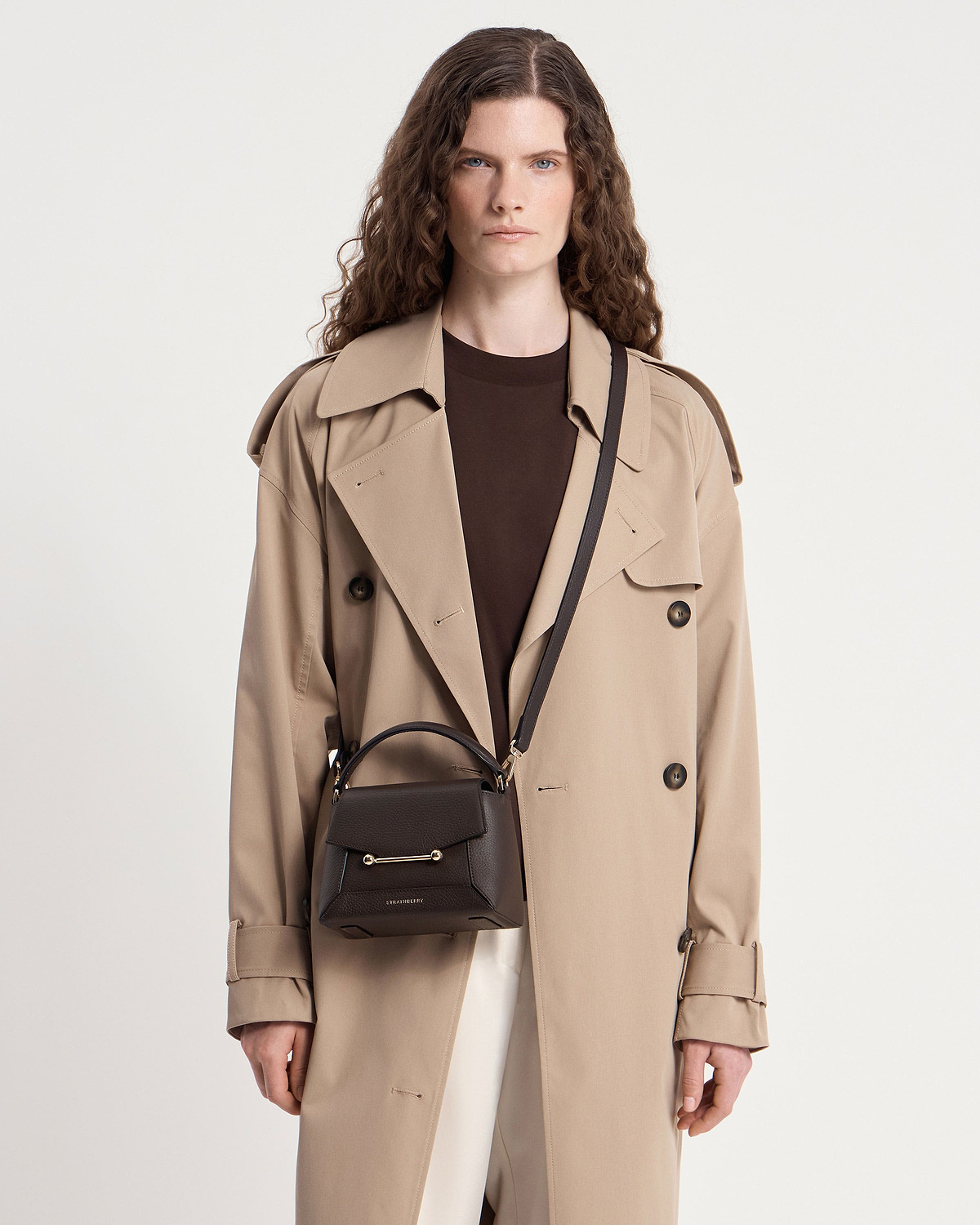 Model wearing trench coat with small Strathberry Mosaic Nano espresso leather top-handle bag.