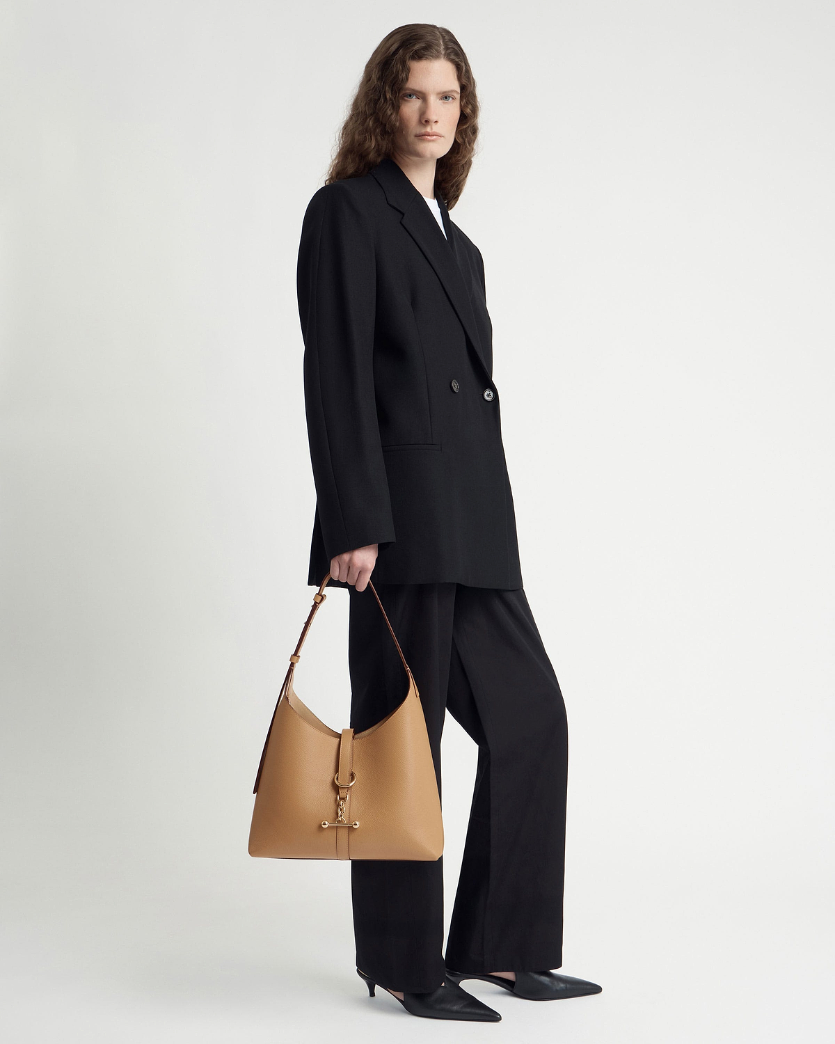 Model in black suit holding caramel Kite Hobo bag by top strap.