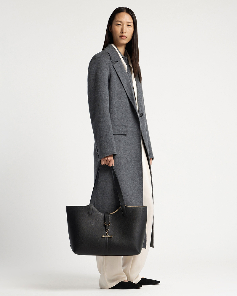 Model in grey coat holding black tote bag with gold hardware by her side.