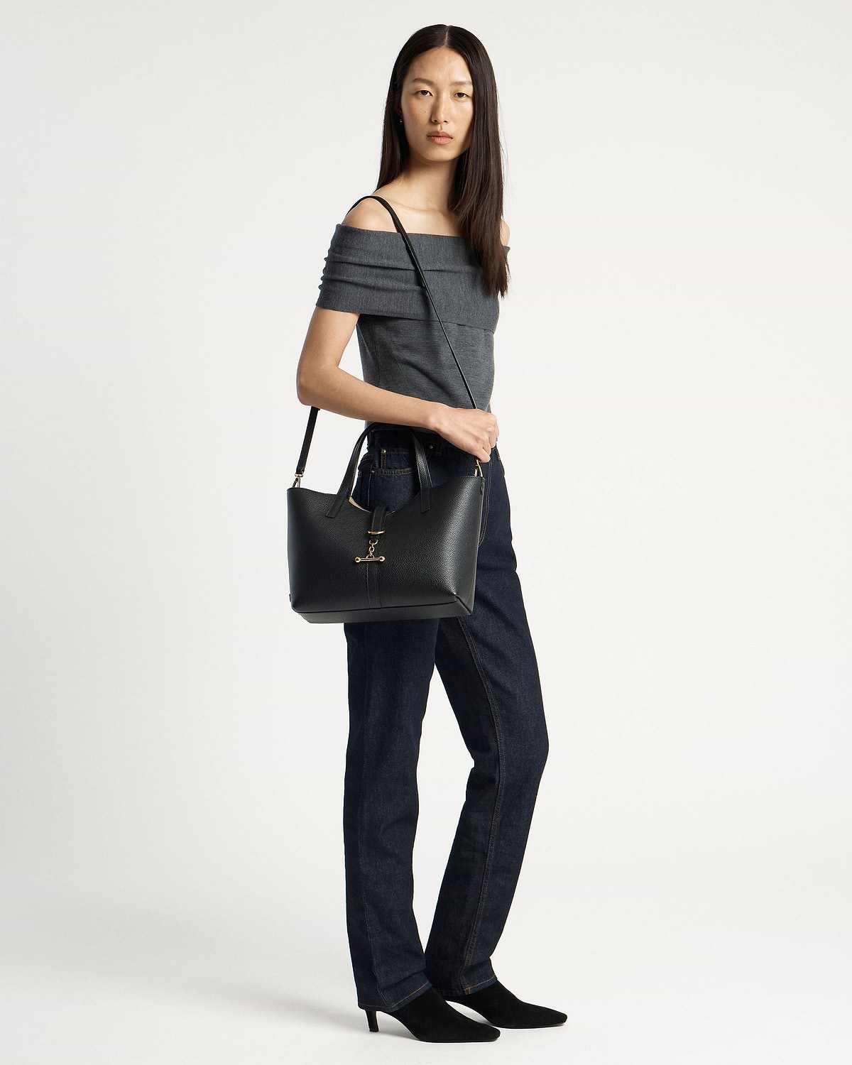 Model wearing grey off-shoulder top and dark jeans carrying black tote bag.
