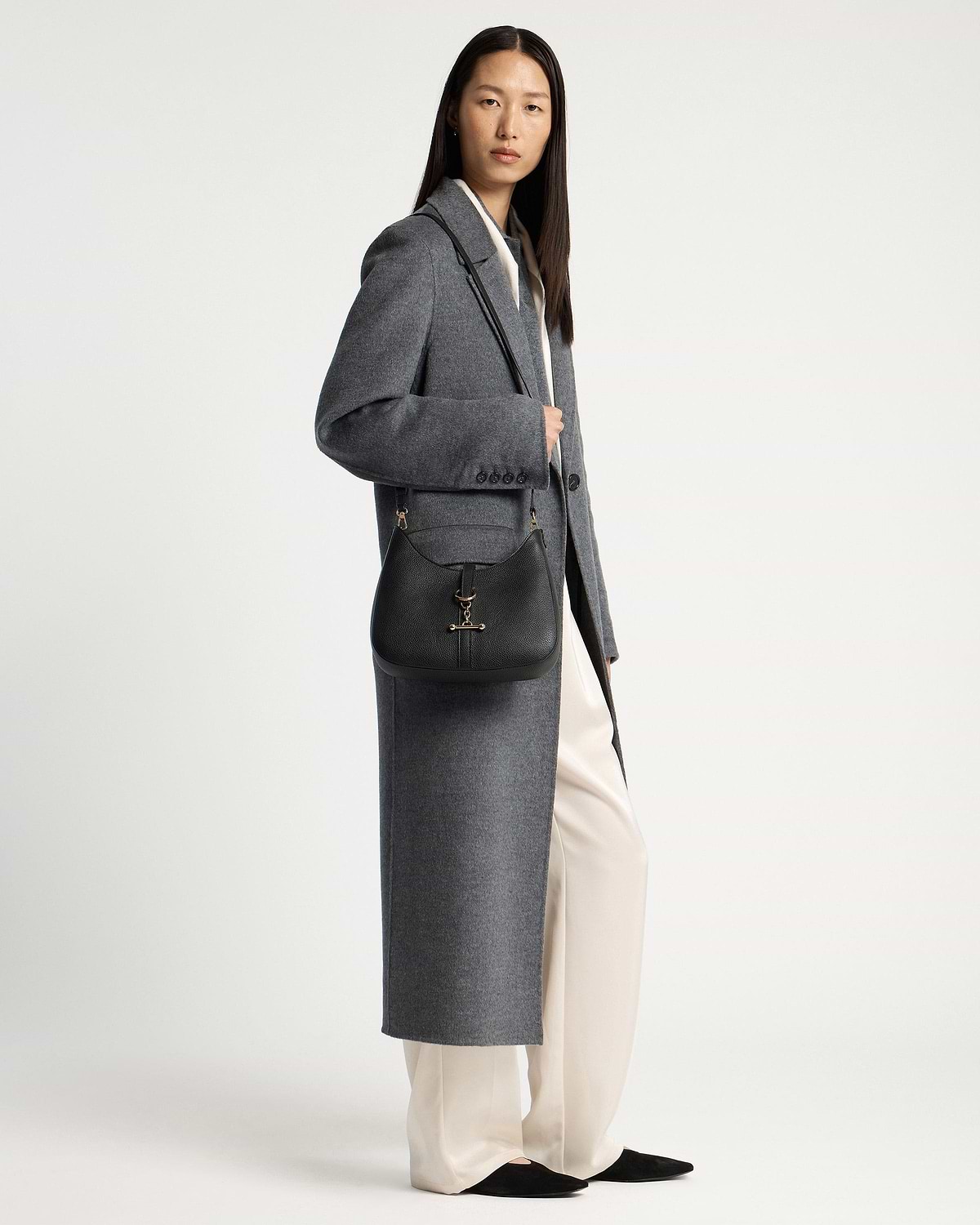 Model wearing long grey coat carrying a black kite sling bag with gold hardware.