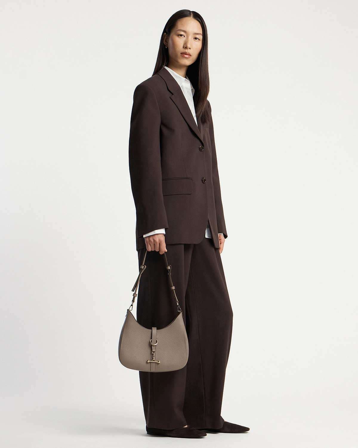 Model holding taupe kite sling bag with gold hardware, wearing dark brown suit.