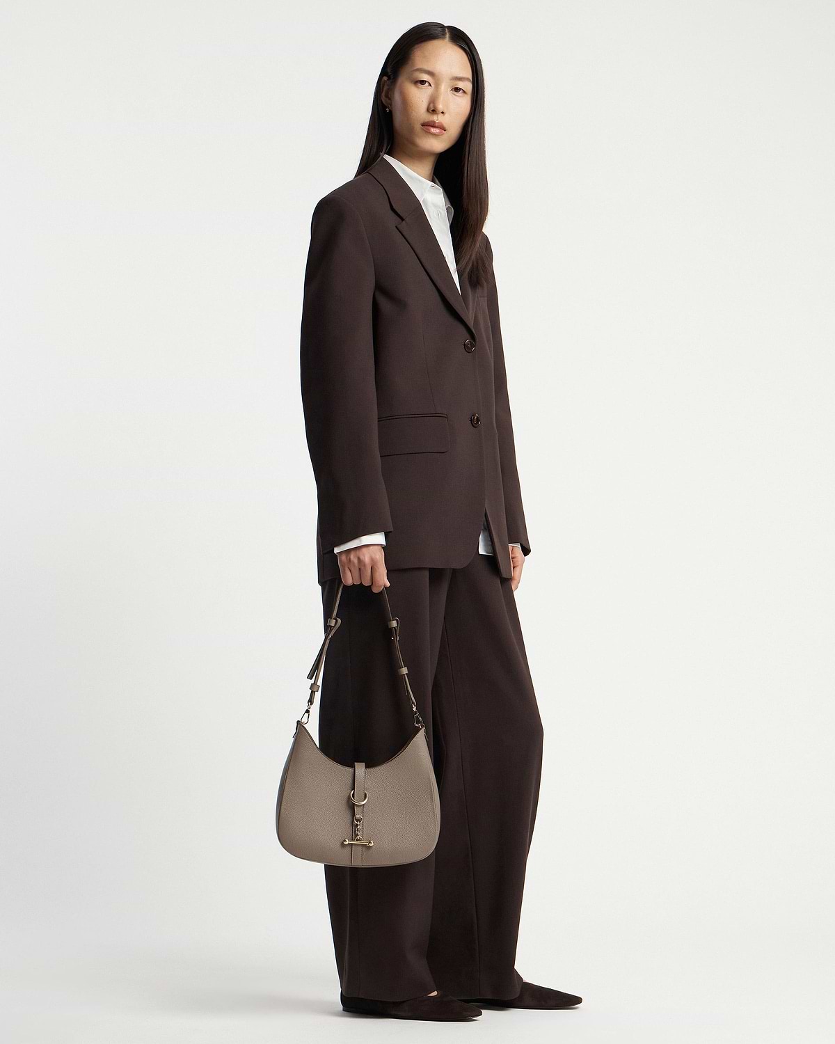 Model holding taupe kite sling bag with gold hardware, wearing dark brown suit.