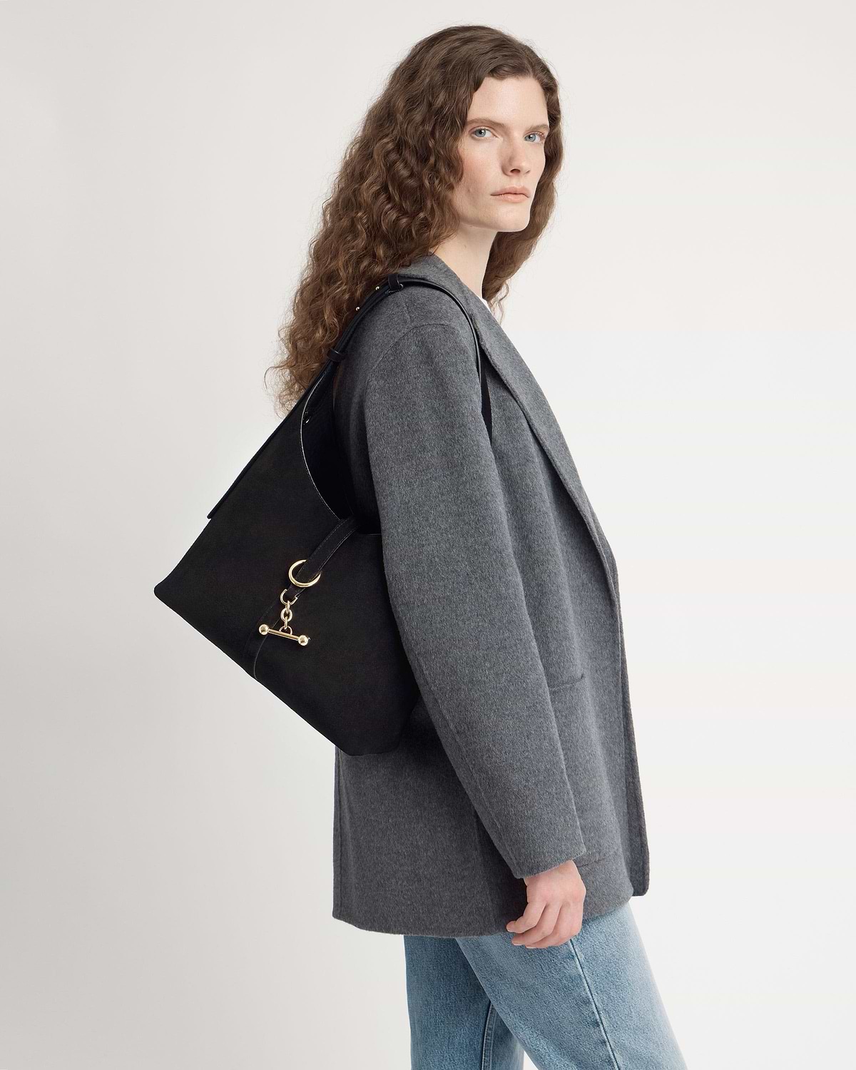 Model in grey coat wearing black suede hobo bag with gold hardware.